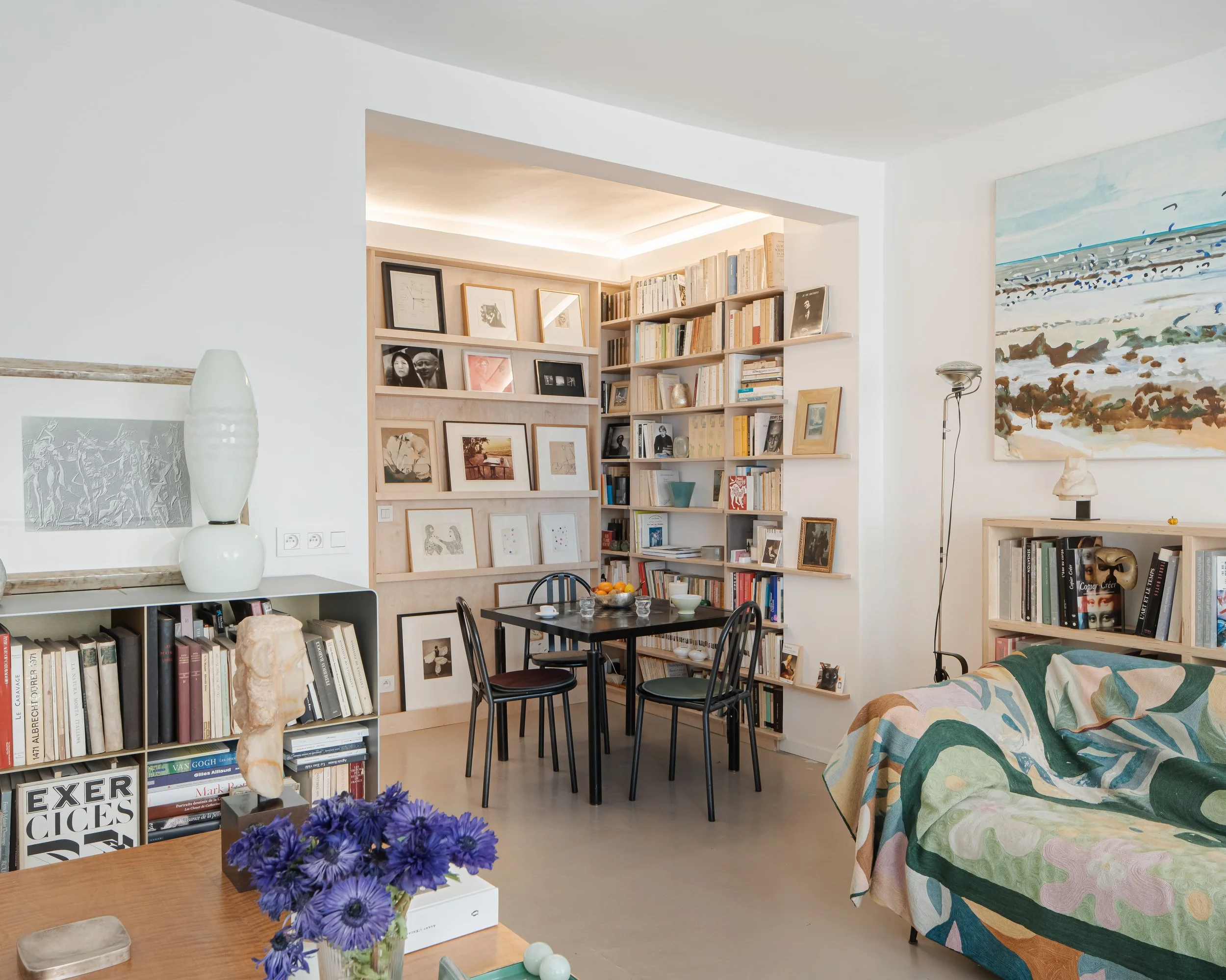 Salon avec bibliothèque, table à manger, canapé recouvert d'une couverture colorée, et diverses œuvres d'art accrochées aux murs.