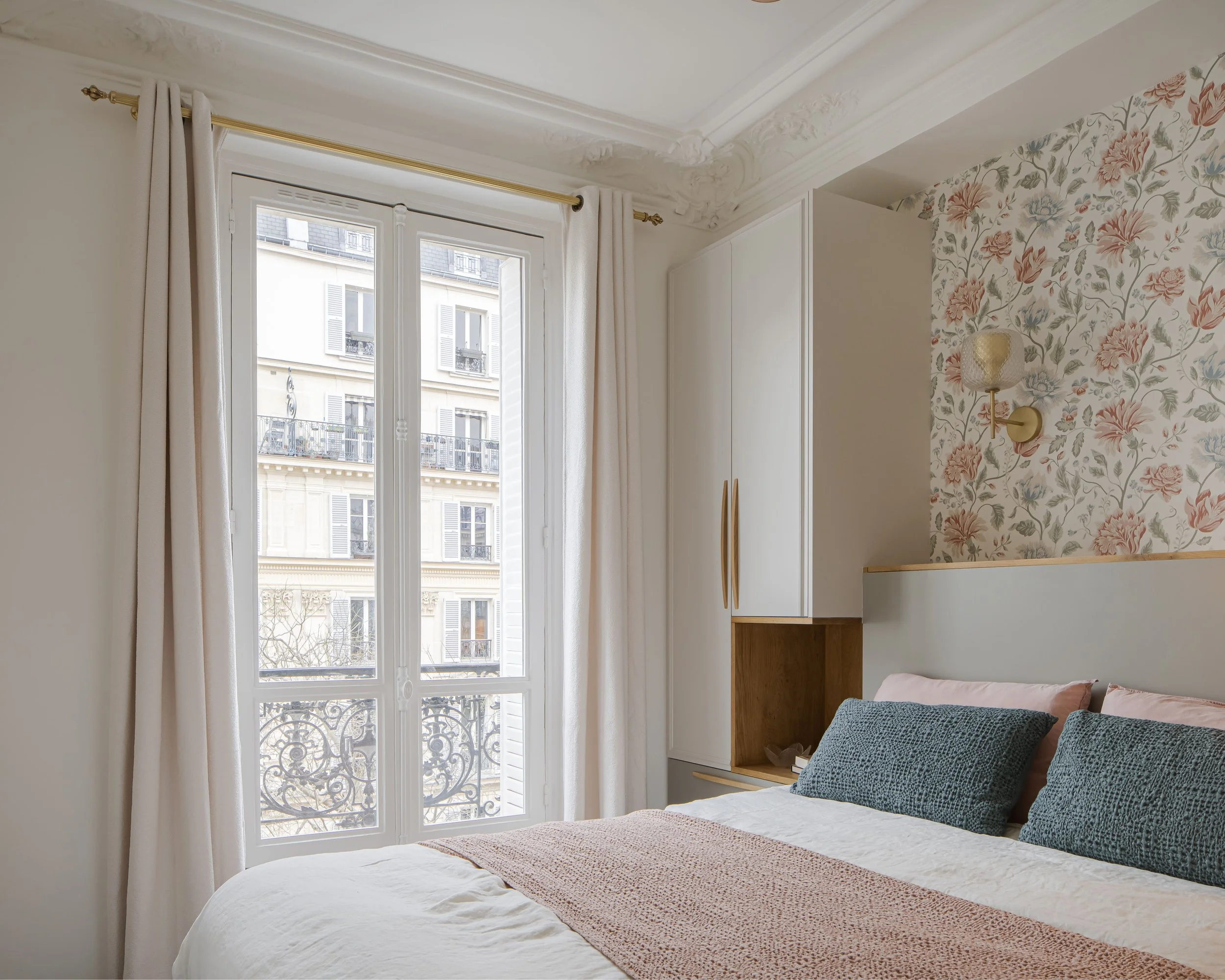 Chambre à coucher avec fenêtre, rideaux blancs, papier peint floral, lit avec oreillers et couvre-lit beige, mur avec appliqué de lampe