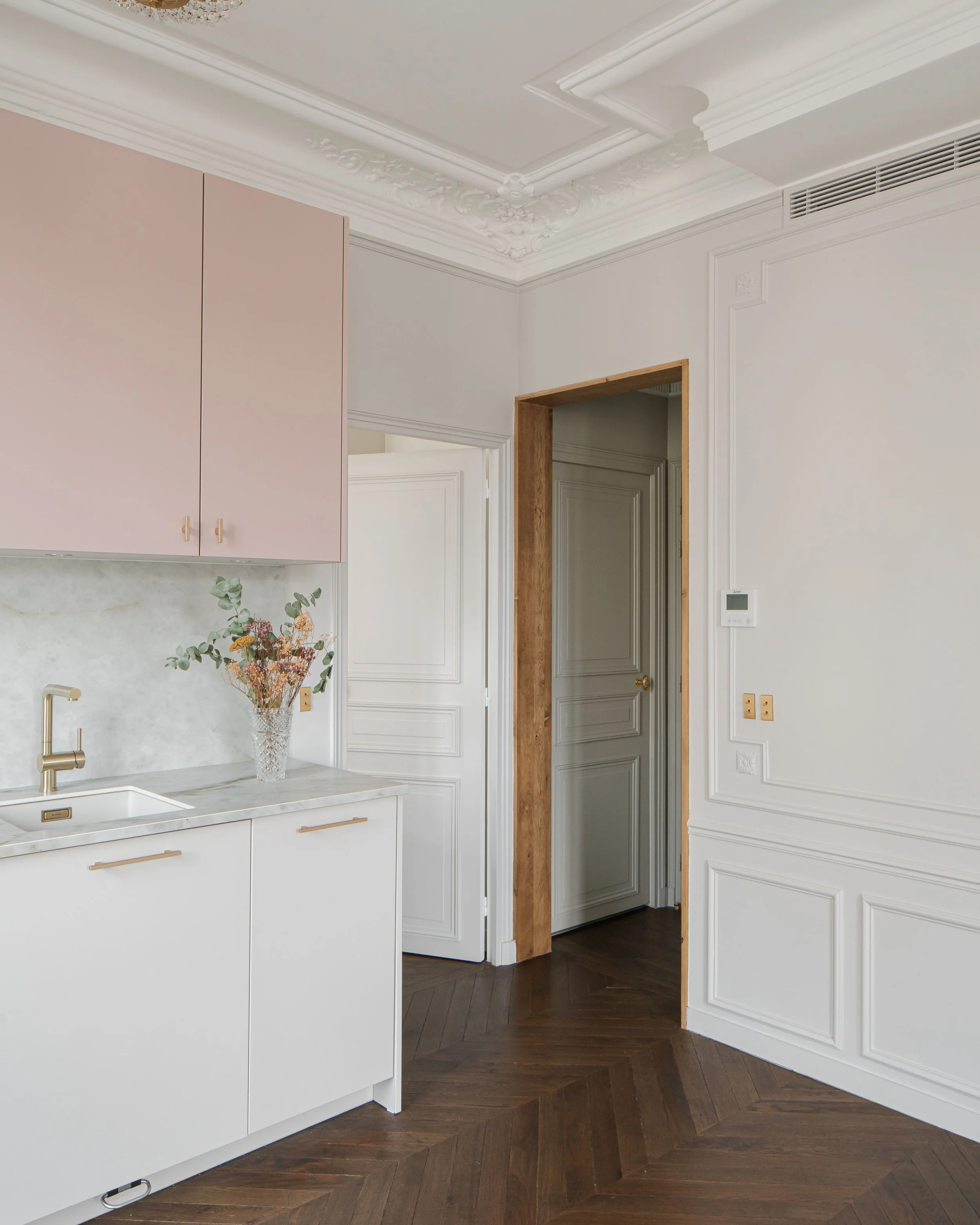 Une cuisine élégante avec des murs blanc cassé, un plan de travail en marbre, des armoires roses, une vase avec des fleurs, un carrelage en bois foncé, et une porte en bois naturel.