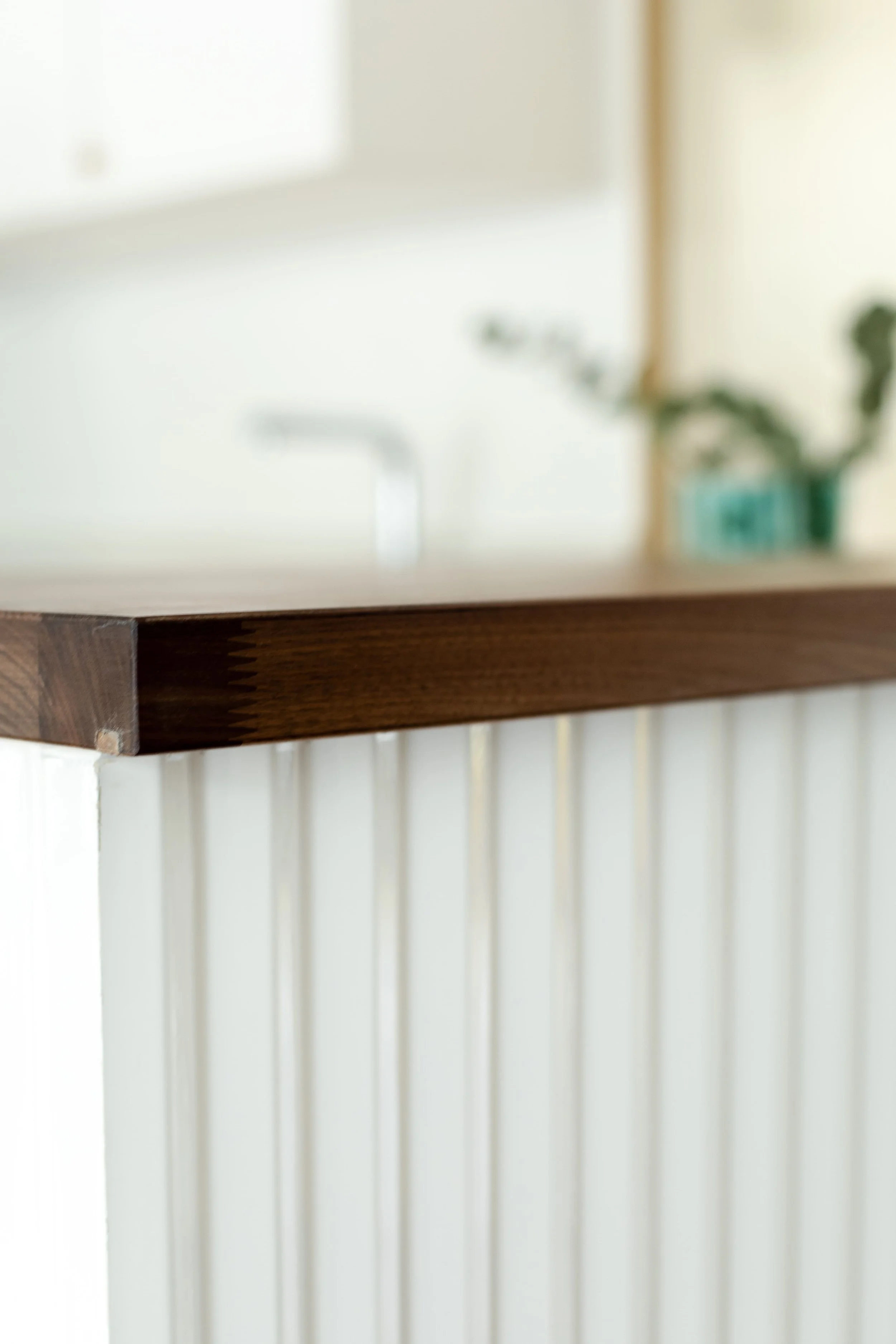 Premier plan d'une surface en bois foncé sur un comptoir en blanc, avec un fond flou montrant un évier et une plante en pot vert