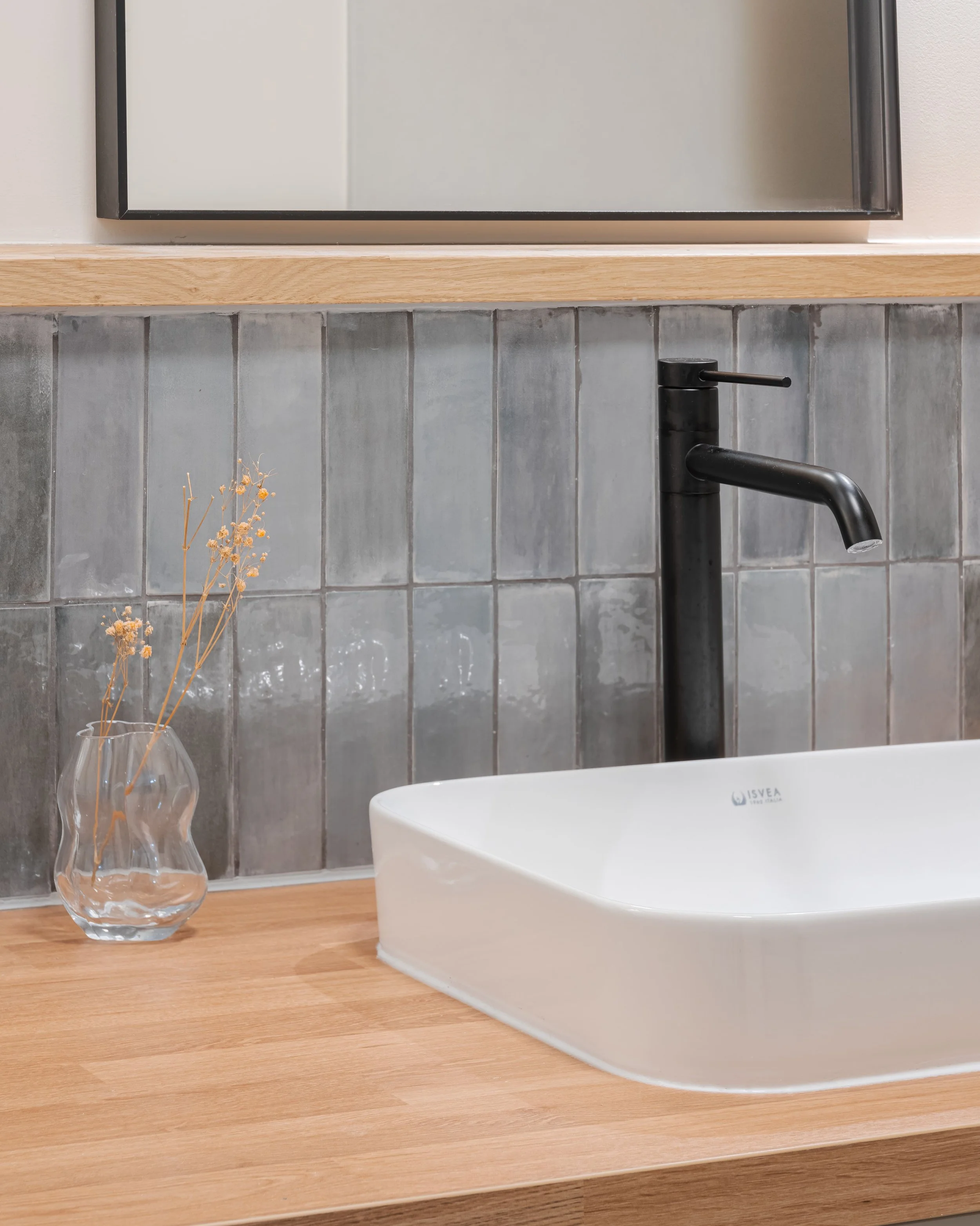 Lavabo en céramique blanc avec robinet noir, miroir au-dessus, vase transparent avec des branches sèches, mur en carreaux gris, plan de travail en bois.