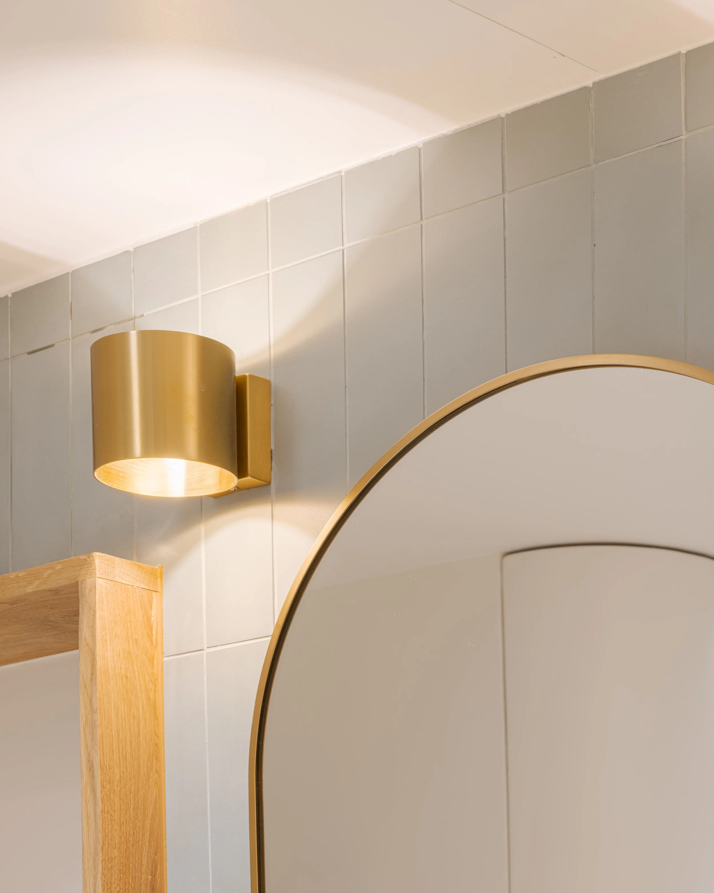 Miroir avec cadre en métal doré dans une salle de bain, mur en carreaux gris clair, applique murale dorée à gauche