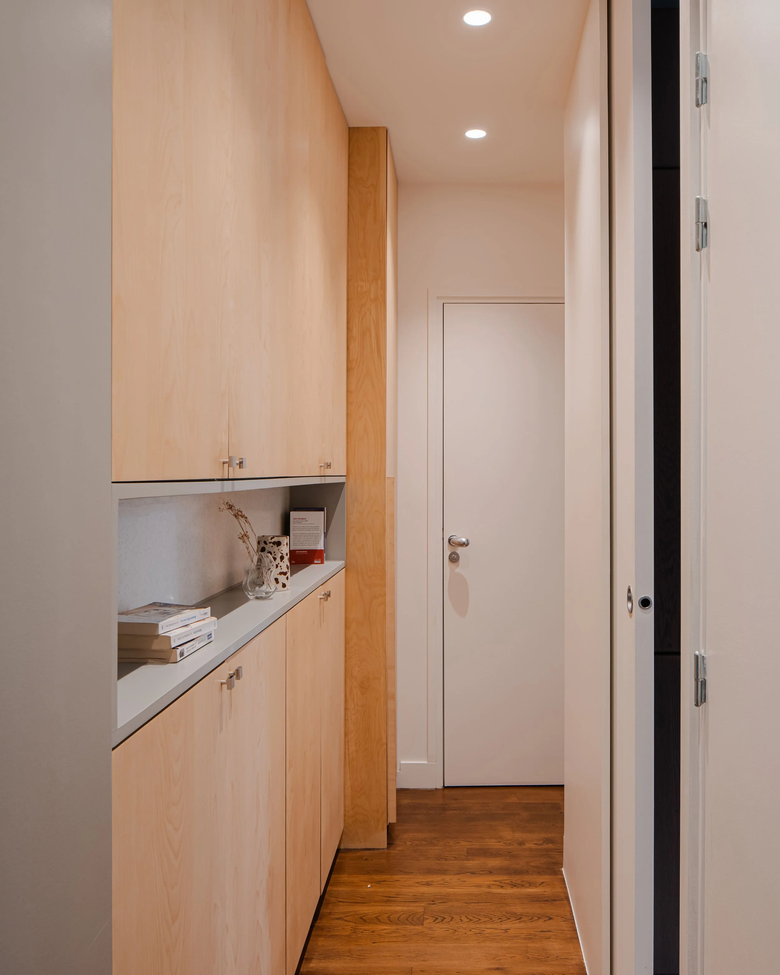 Entrée d'un couloir avec des armoires en bois clair, une porte blanche au fond, un sol en bois et un mur blanc.