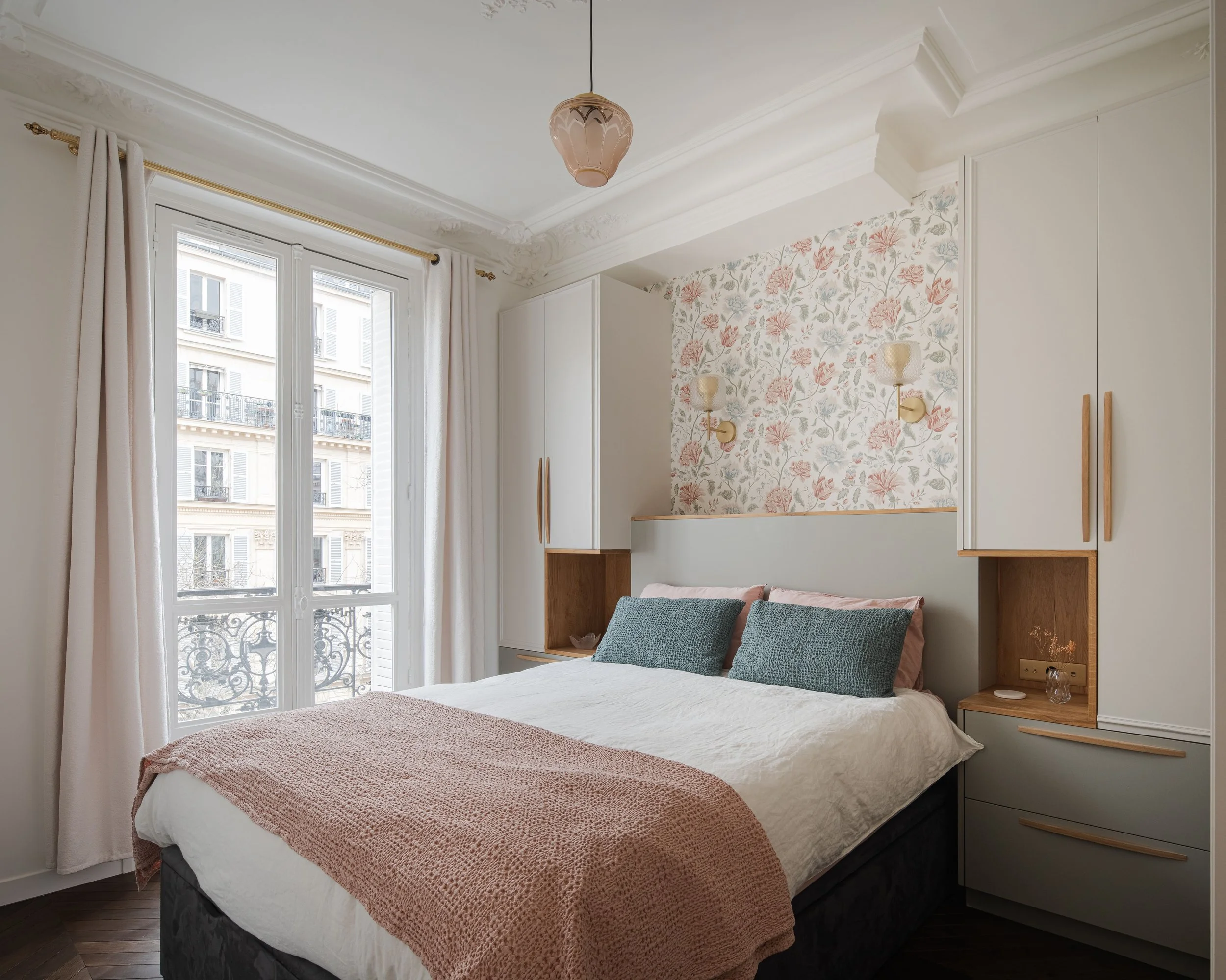 Chambre avec lit double, accents de rose et vert, armoires blanches encadrant le lit, sur fond de papier peint floral pastel, près d'une fenêtre avec rideaux blancs et balcon en fer forgé.
