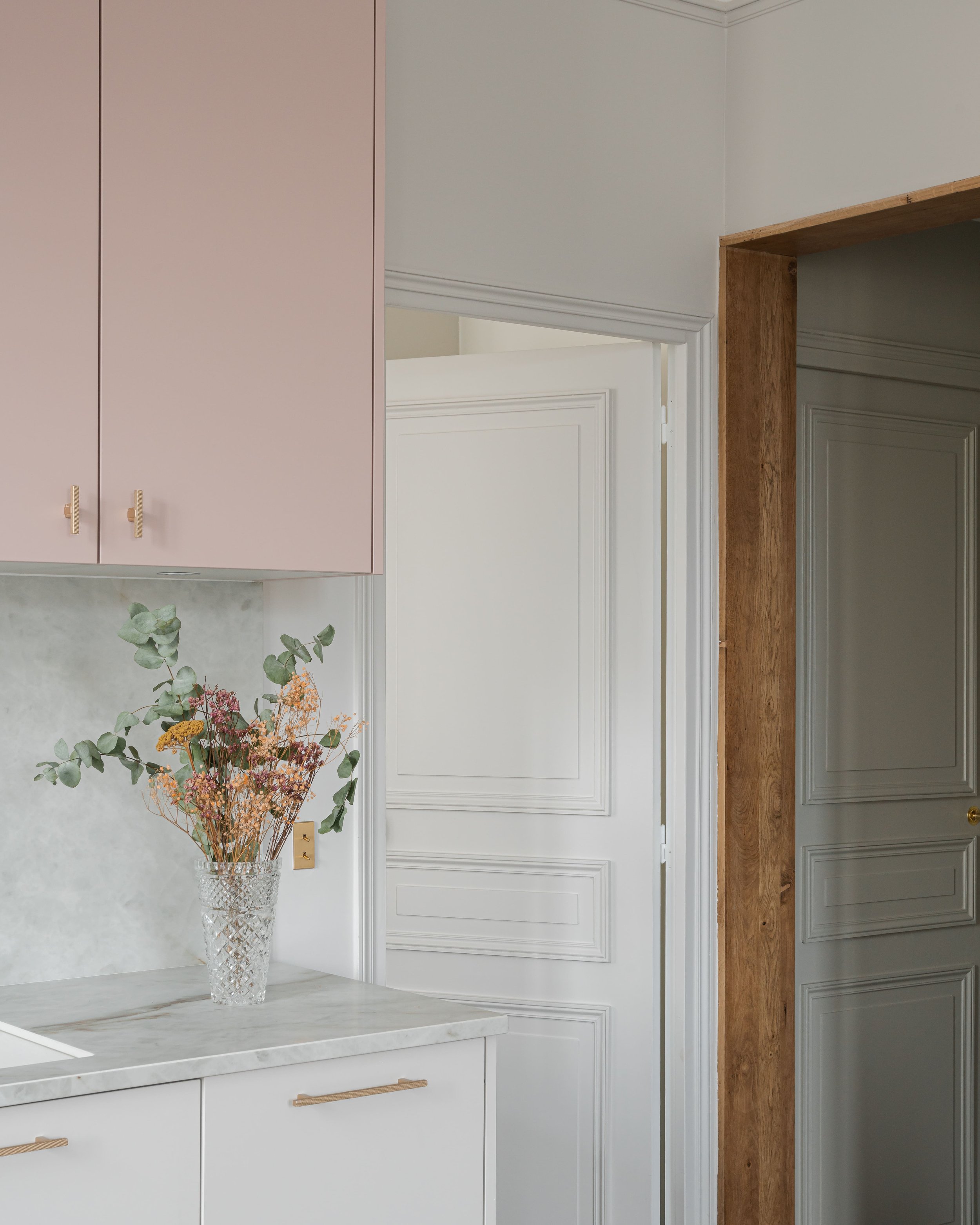Intérieur de cuisine avec vase de fleurs, armoires roses, portes blanches avec moulures, cadre en bois autour de la porte.