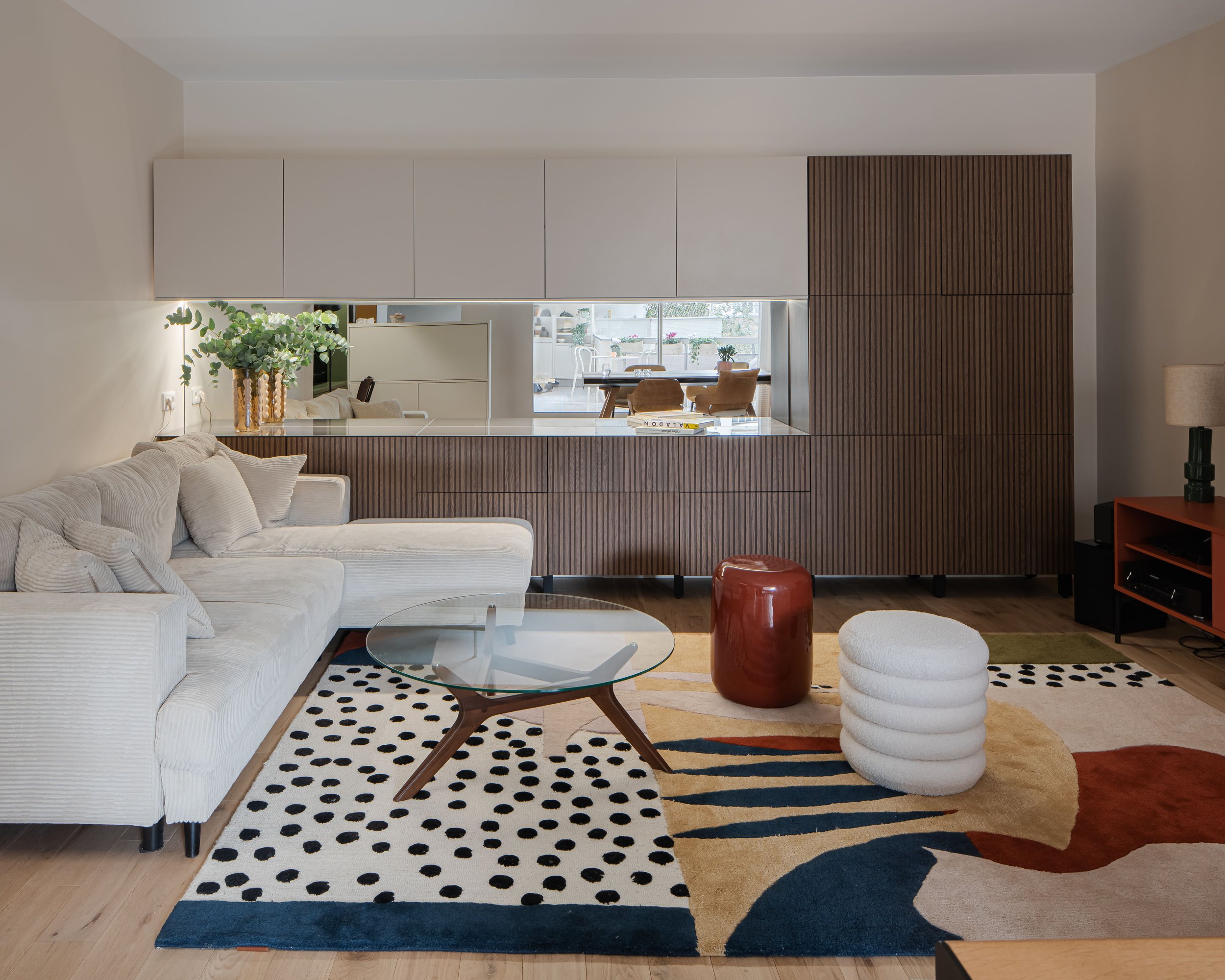 Salon moderne avec canapé beige, tapis coloré, table en verre, décoration élégante, bibliothèque en bois, et vue sur une véranda