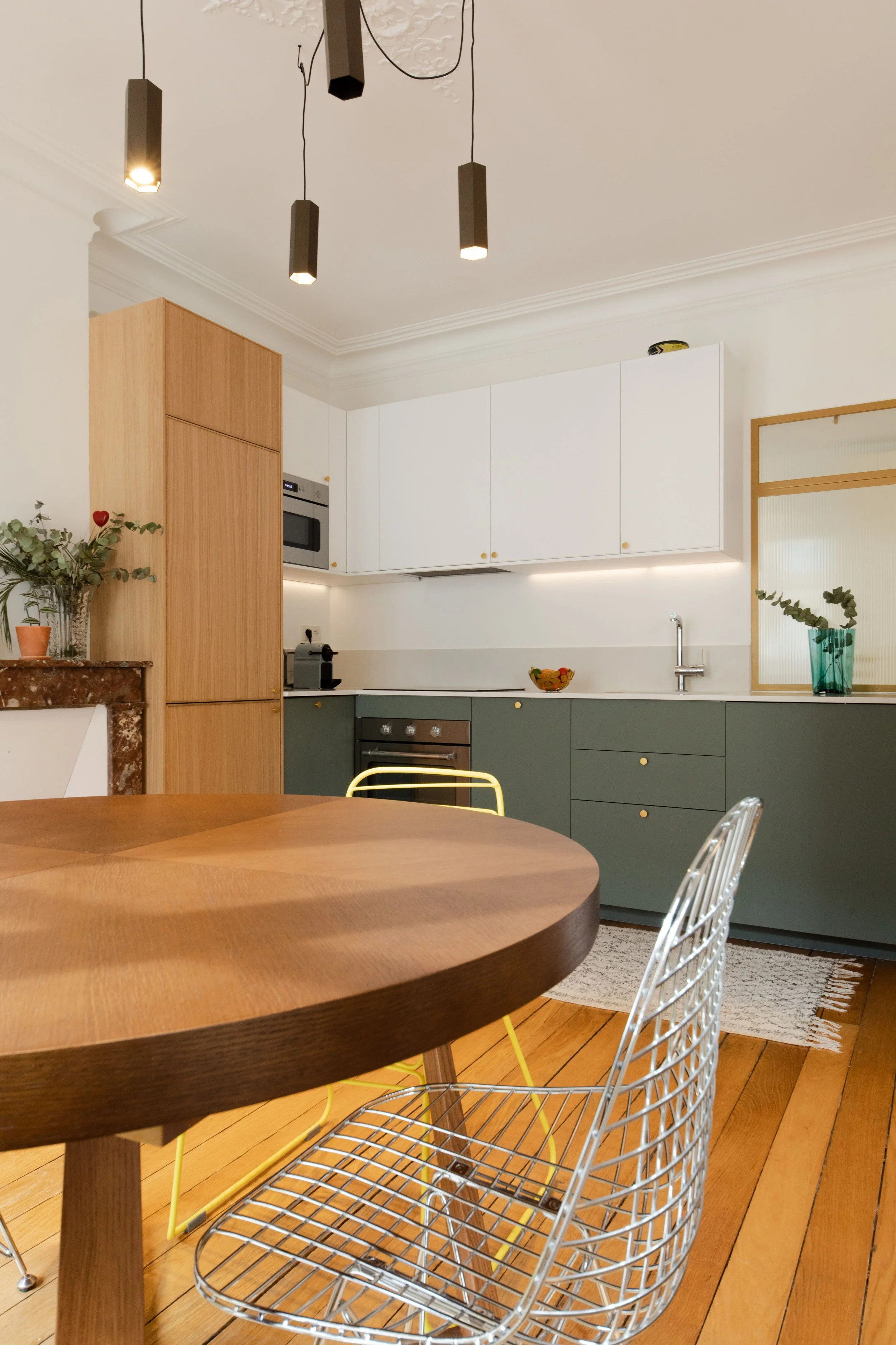 Cuisine moderne avec un mur blanc, des armoires blanches et vertes, un table en bois rond, des chaises en fil métallique, une lampe suspendue noire, et des plantes décoratives.