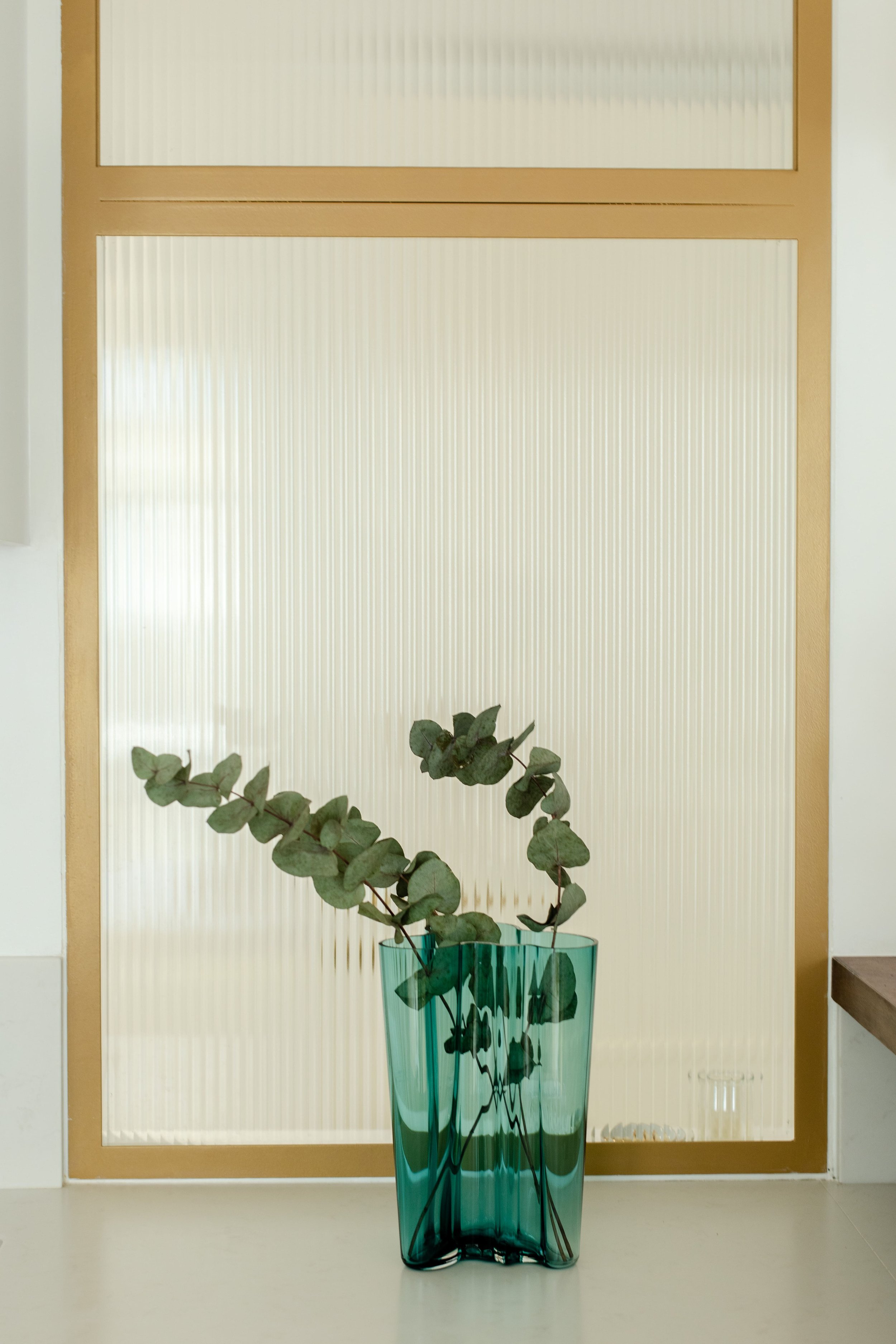 Un vase en verre vert contenant des branches de eucalyptus, placé devant une porte en verre translucide avec cadre en bois.