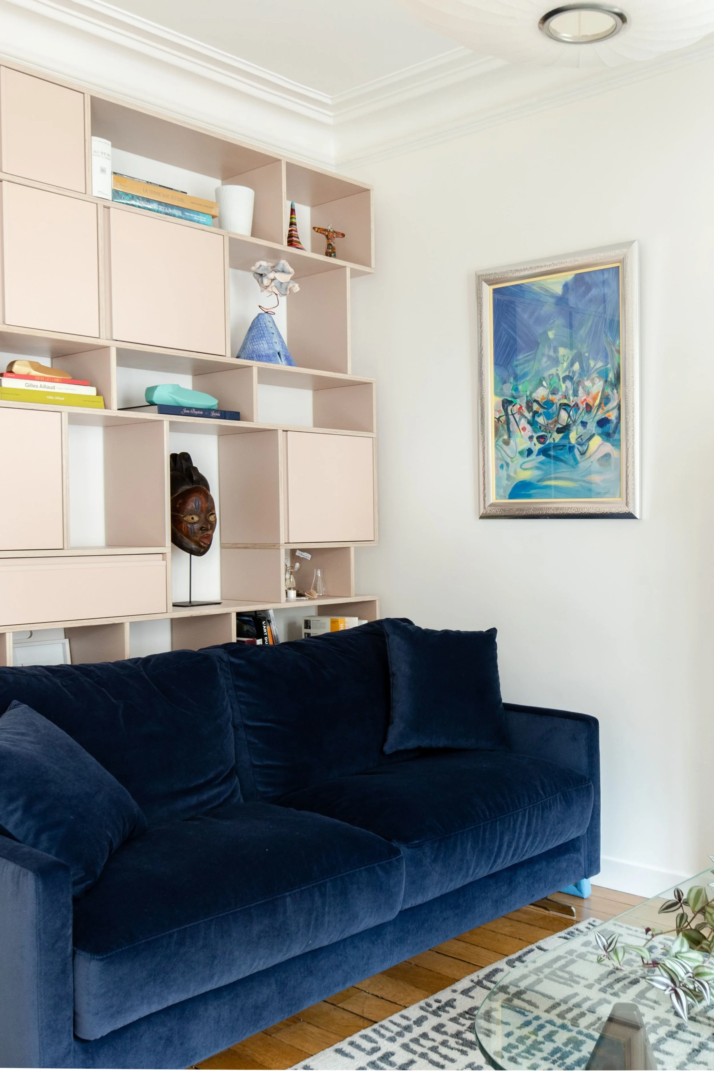 Salon avec un canapé en velours bleu foncé, étagère moderne avec des livres, une sculpture et un masque africain. Mur blanc avec peinture abstraite.