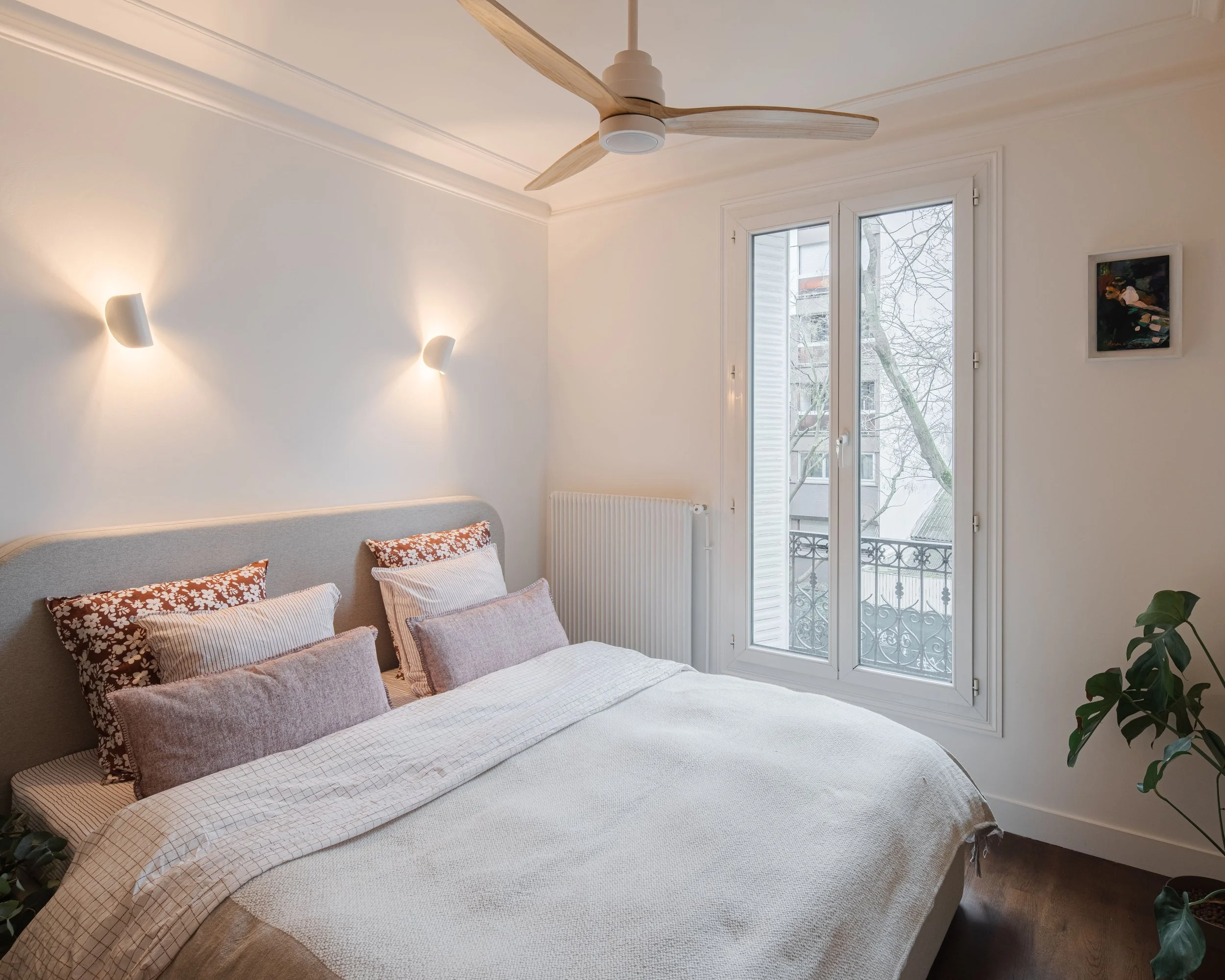 Chambre lumineuse avec lit double, plusieurs oreillers, fenêtre donnant sur un balcon avec des arbres nus, et un plafond avec ventilateur en bois.