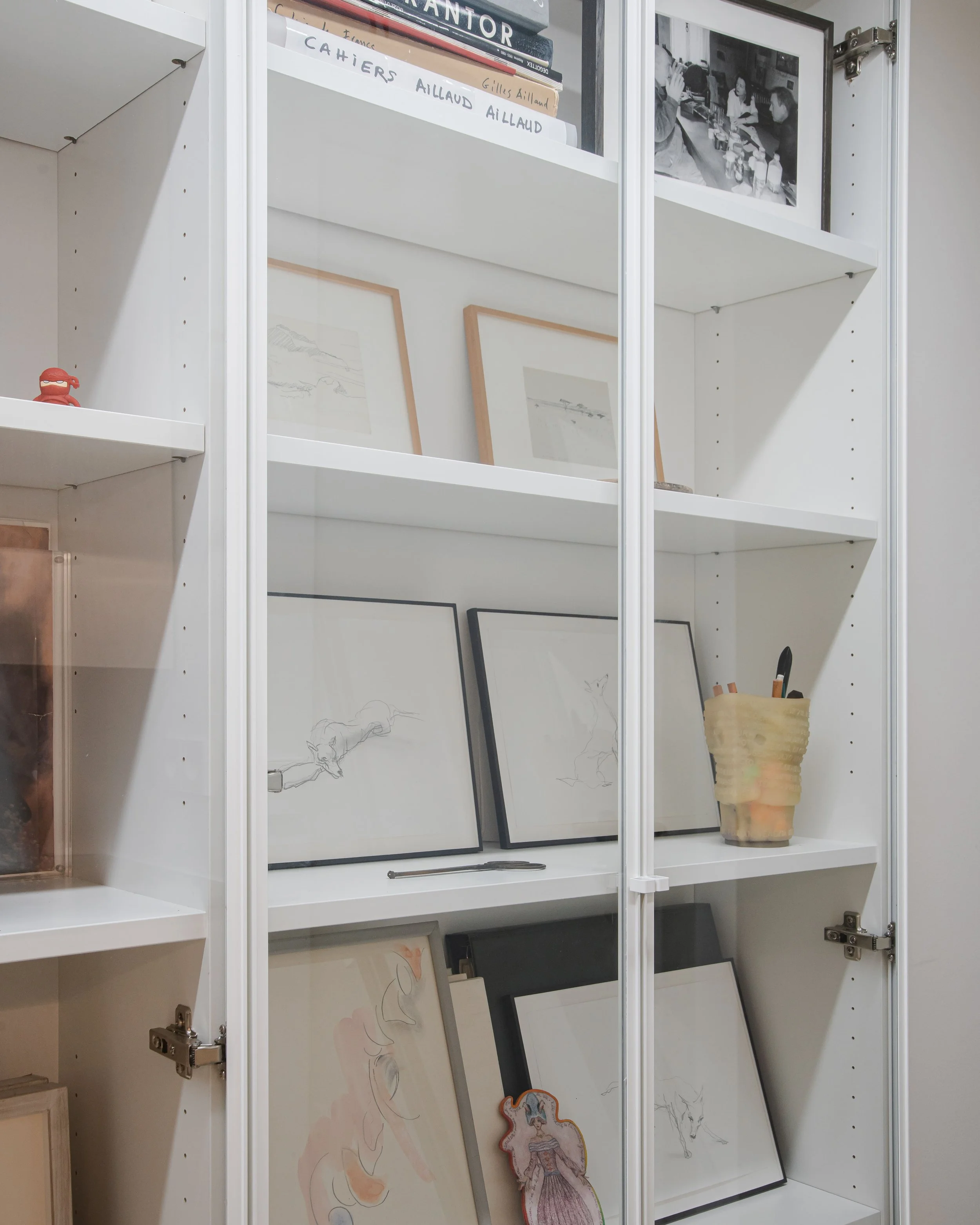 Une étagère blanche contenant des glaces, des livres, des dessins encadrés, une photo en noir et blanc, et un pot avec des stylos.