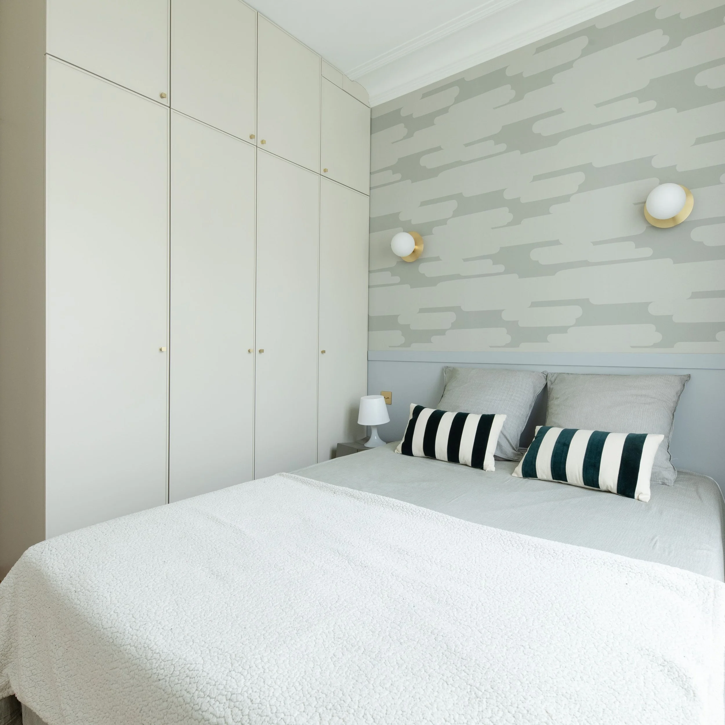 Chambre à coucher avec un lit, deux coussins rayés noir et blanc, deux lampes de chevets, un mur décoratif avec un motif abstrait, un placard blanc avec des poignées dorées, une lampe murale et un mur de couleur neutre.