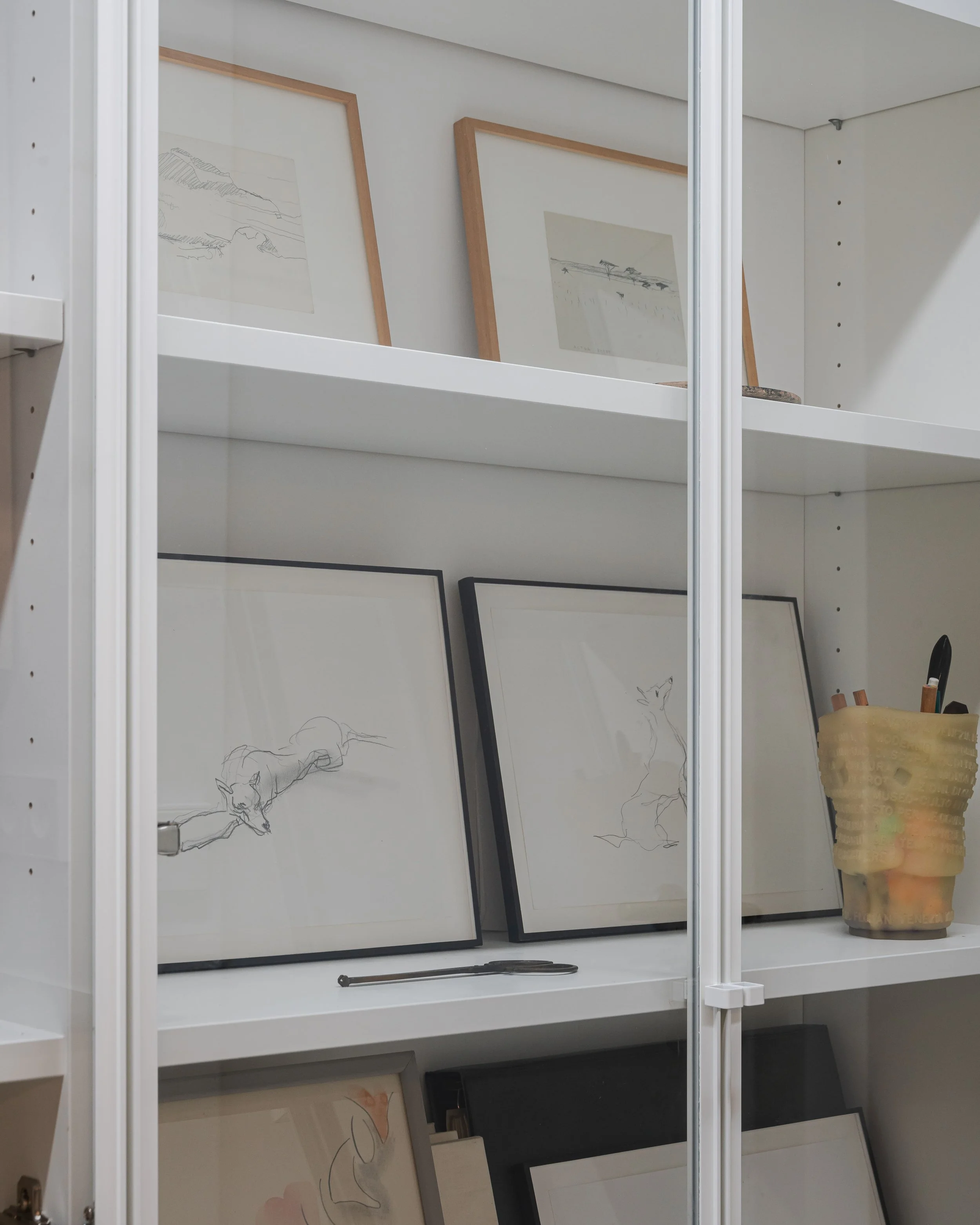 Étageroire avec plusieurs cadres de dessins de peintures ou illustrations, dont certains sont esquissés avec des lignes minimalistes d'animaux, ainsi qu'un contenant avec des pinceaux et un ciseau.