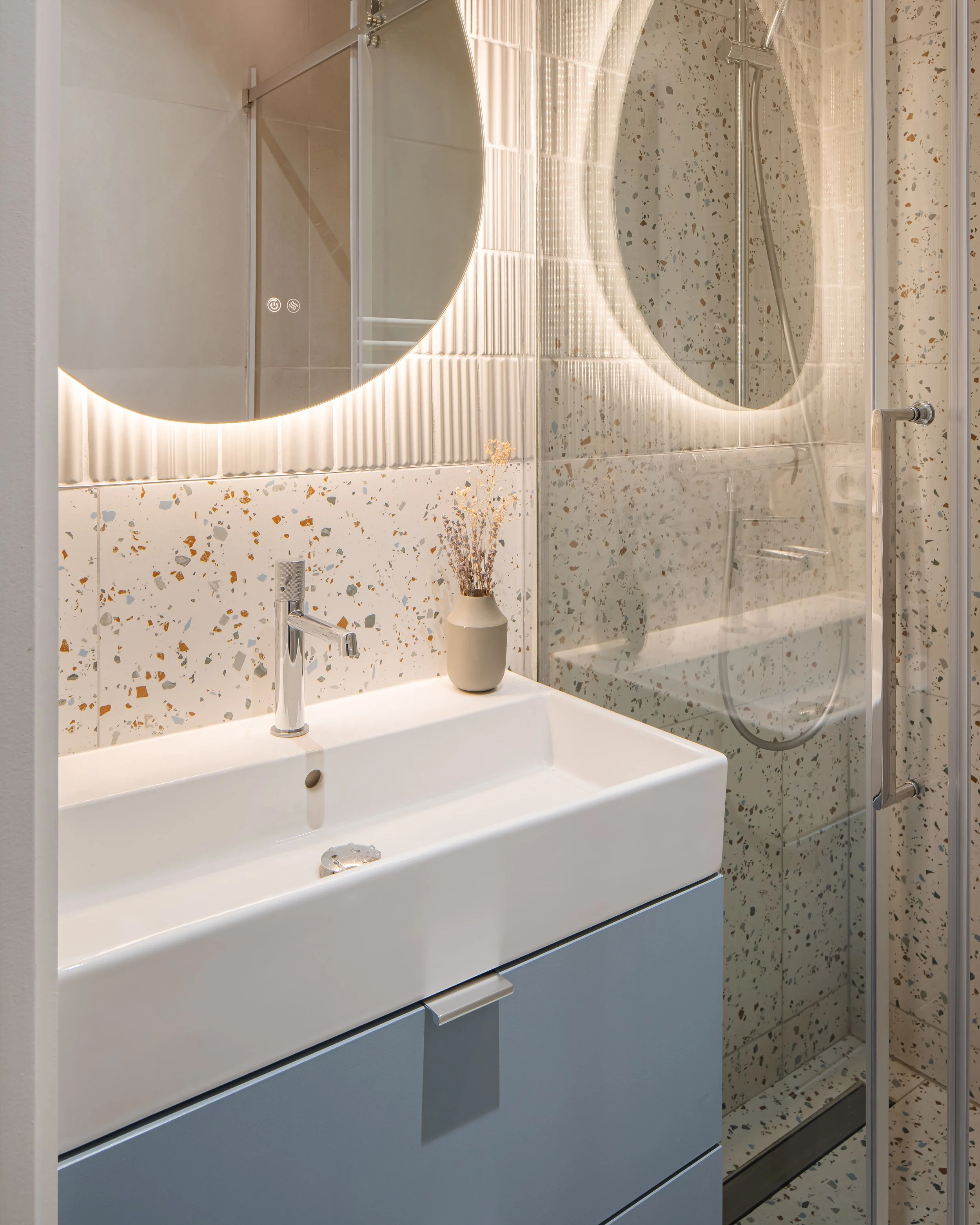Vue d'une salle de bain moderne avec un lavabo blanc sur meuble gris, un miroir rond avec éclairage intégré et une cabine de douche avec porte en verre, carrelages à motifs terrazzo.