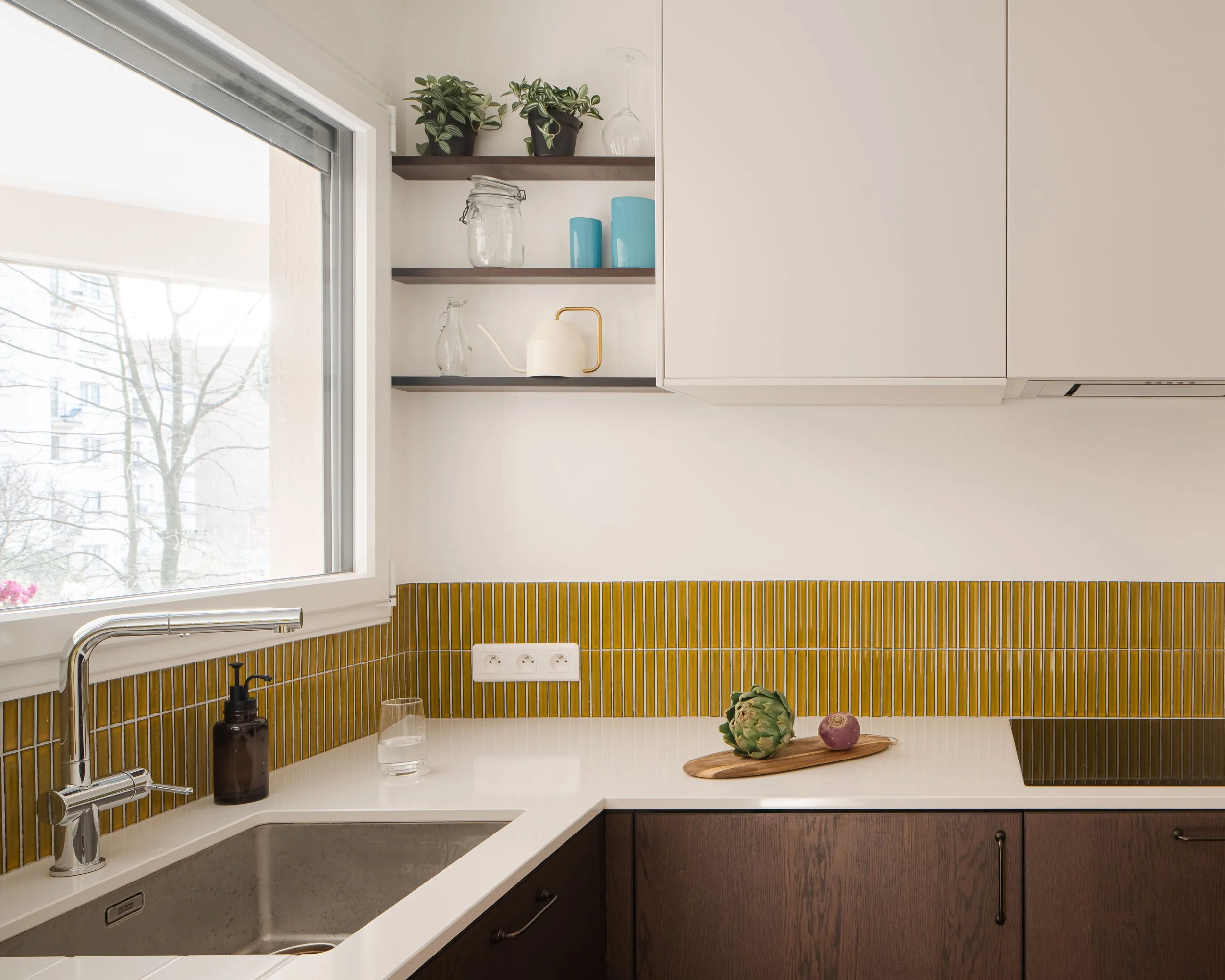 Cuisine moderne avec fenêtre, lavabo, plantes en pot, étagères décoratives, et comptoir avec artichaut et oignon.