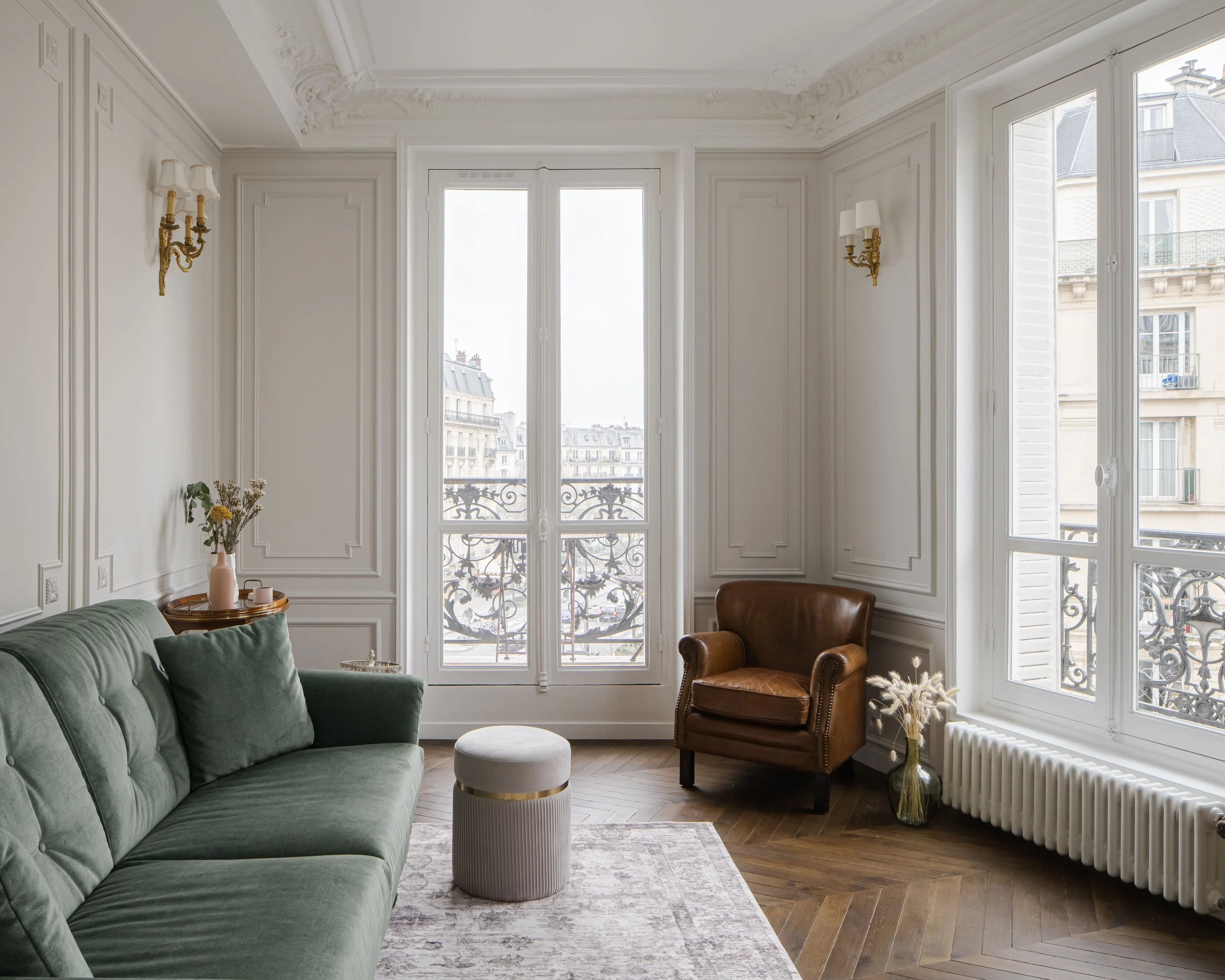 Salon orné de moulures blanches avec deux grands fenêtres, canapé vert, fauteuil en cuir marron, vases avec fleurs, tapis clair, parquet en bois en chevron, deco classique