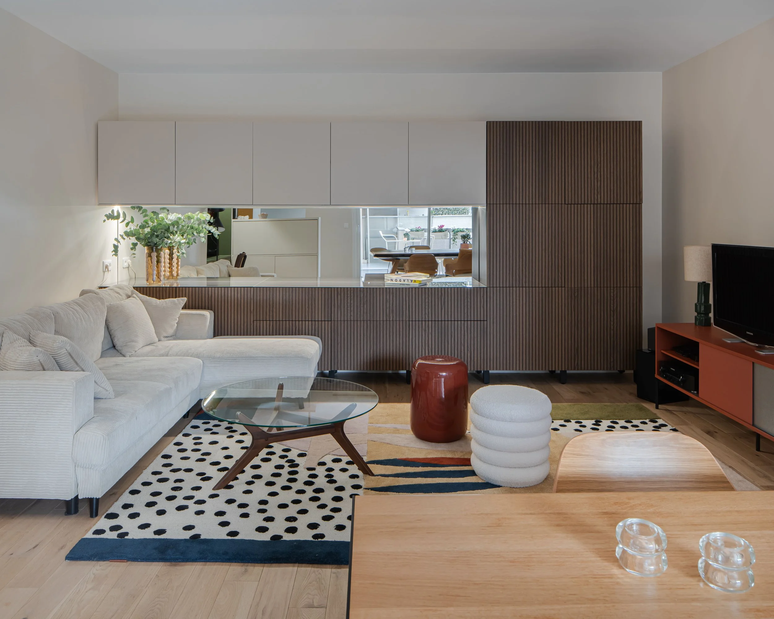 Salon moderne avec canapé beige, tapis à pois noirs et blancs, table en verre, coussin blanc, meuble TV orange, décoration artistique, cuisine derrière avec matériaux en bois, plantes, et fenêtres laissant entrer la lumière.
