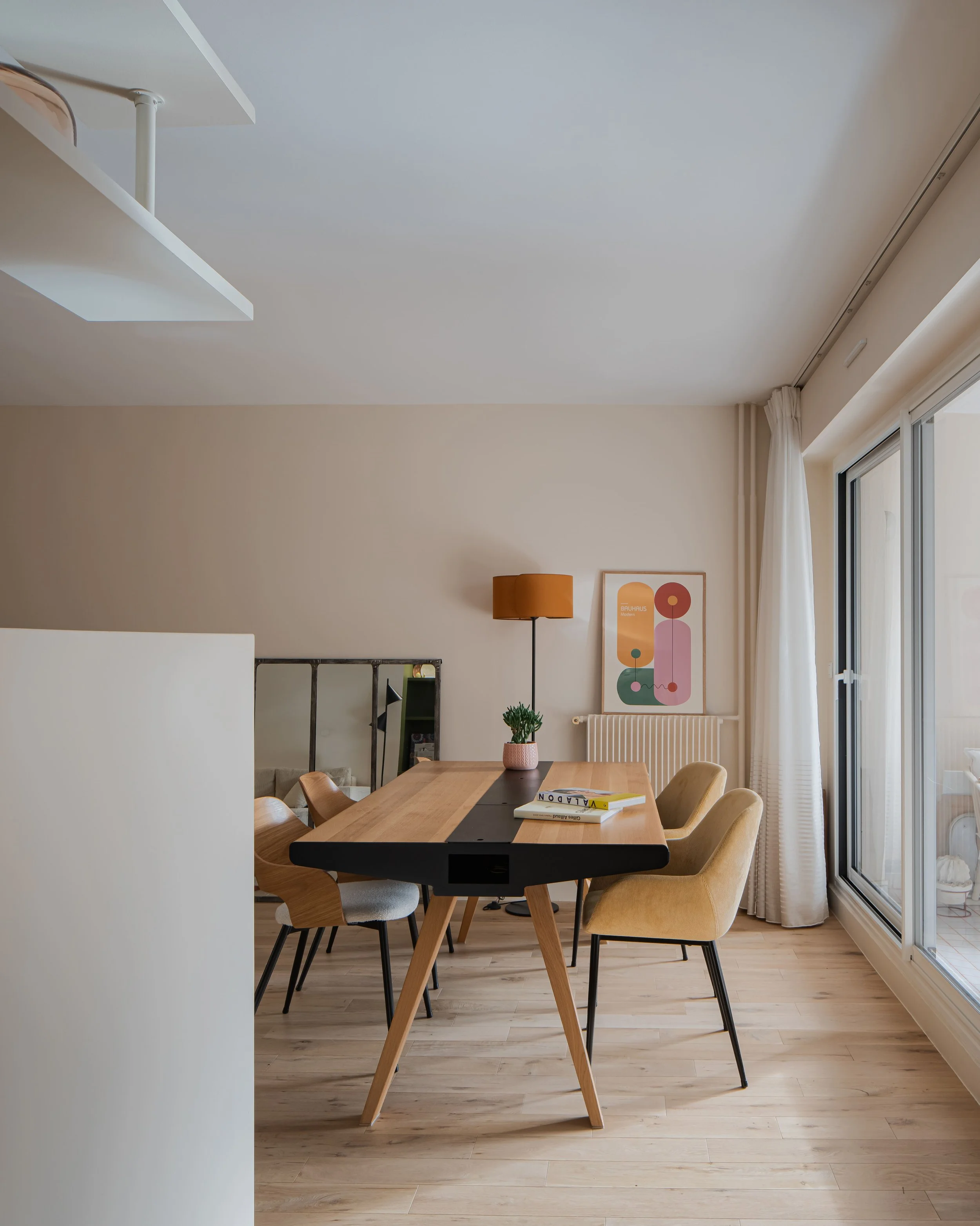 Salle à manger moderne avec un grand tableau graphique sur le mur, une table en bois avec des chaises beige et un lampadaire orange à côté d'une fenêtre.
