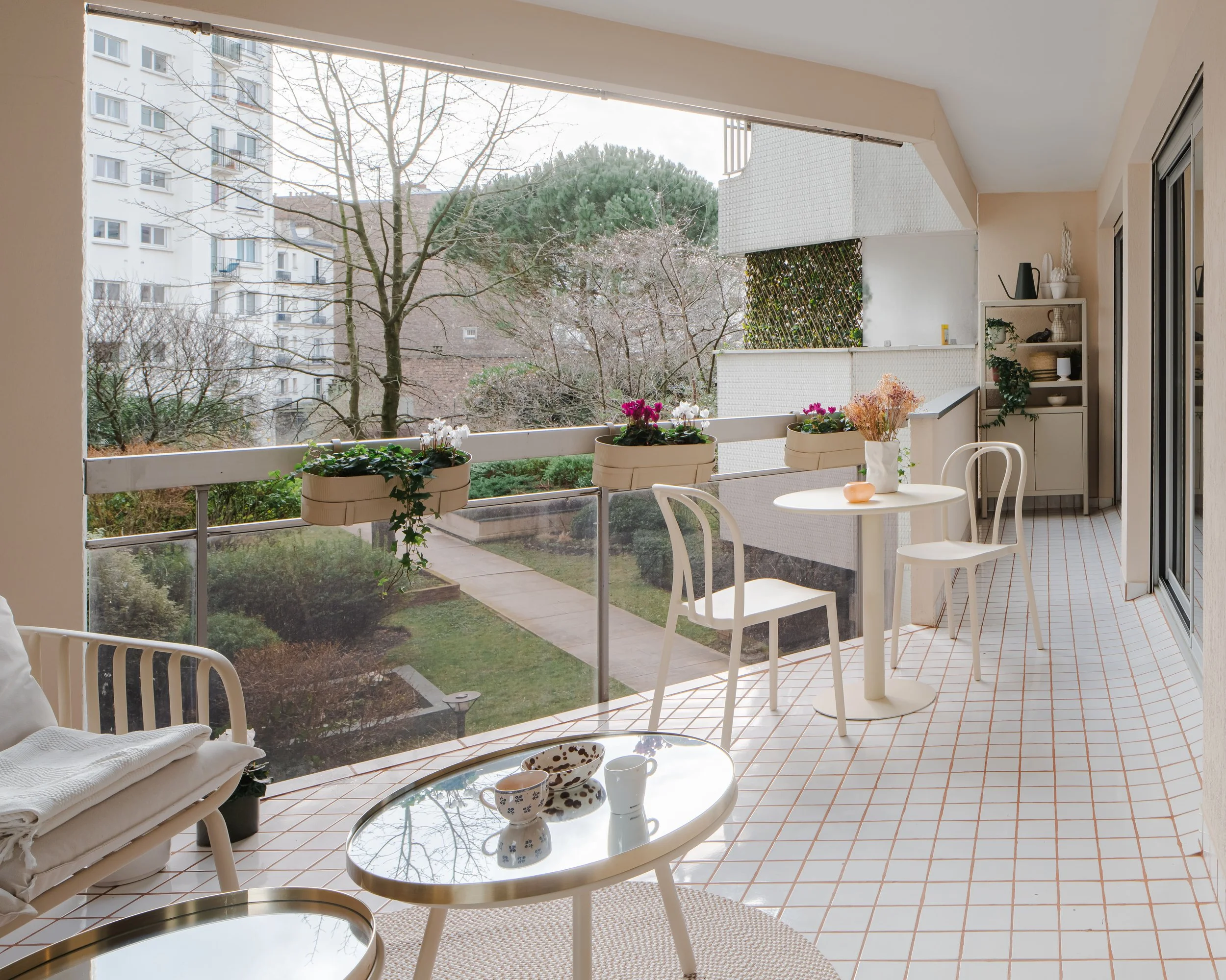 Balcon avec mobilier blanc, plantes en pot, vue sur espace vert et immeubles résidentiels, sol en carreaux de terre cuite