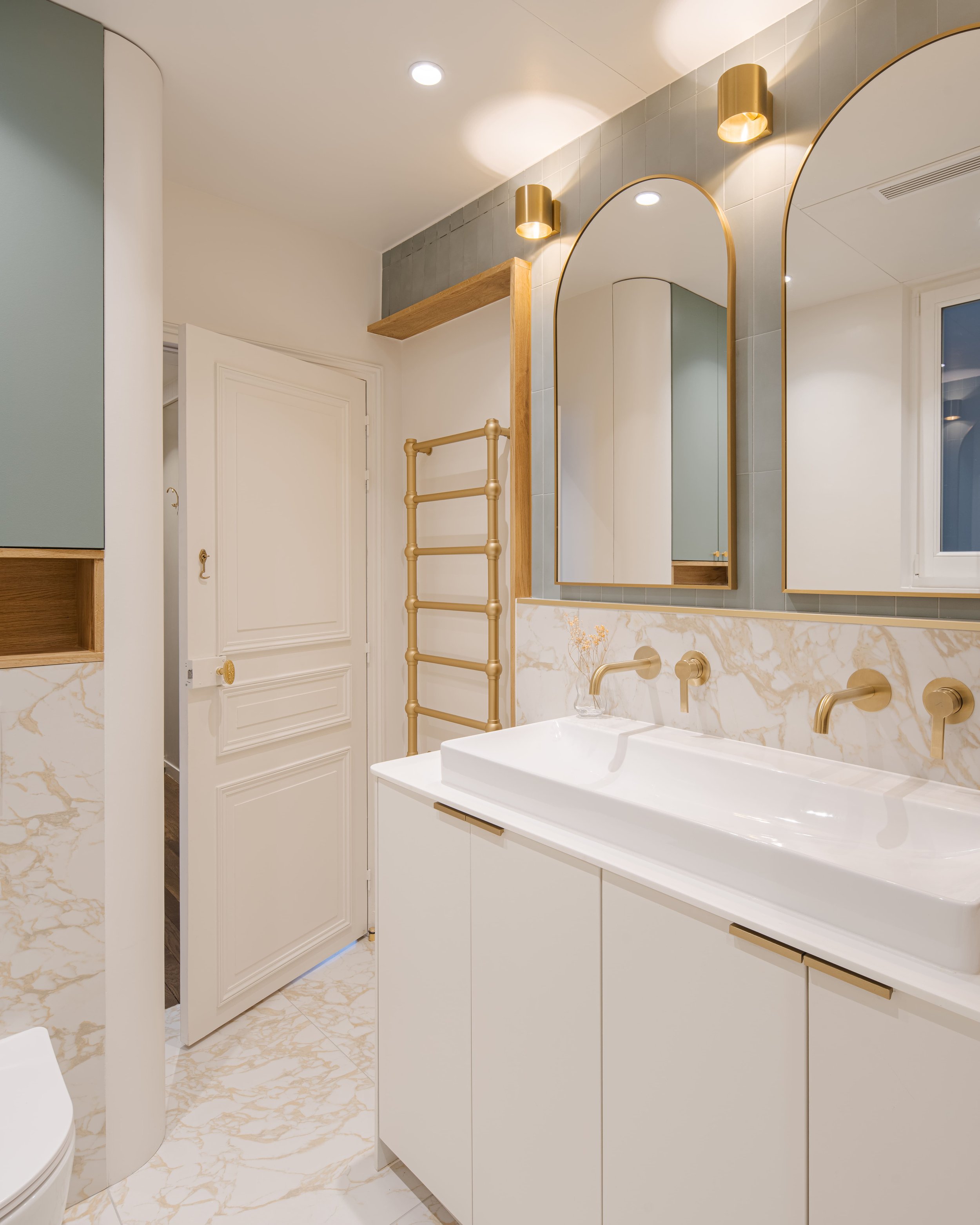 Salle de bain moderne avec double miroir en arc, lavabo blanc avec robinets en or, murs en carrelage gris et blanc, portée de lavabo blanche, étagère en bois, radiateur en forme de barre pour serviettes en or, éclairage encastré au plafond, fenêtre.