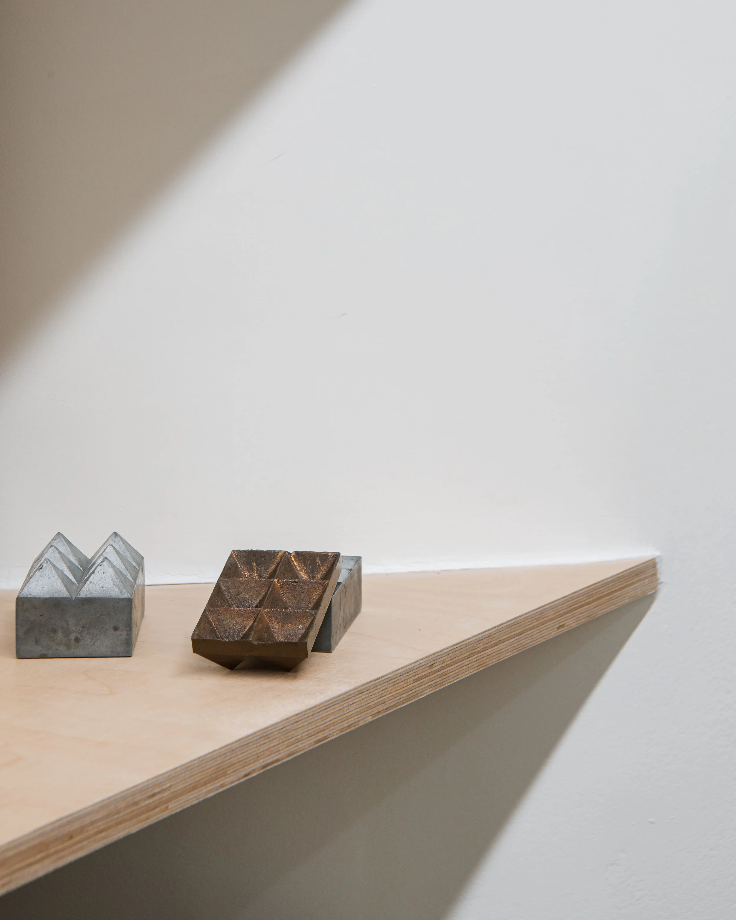 Deux objets en métal, un en forme de montagnes et l'autre en forme de plaque divisée en neuf sections, placés sur une étagère en bois contre un mur blanc.