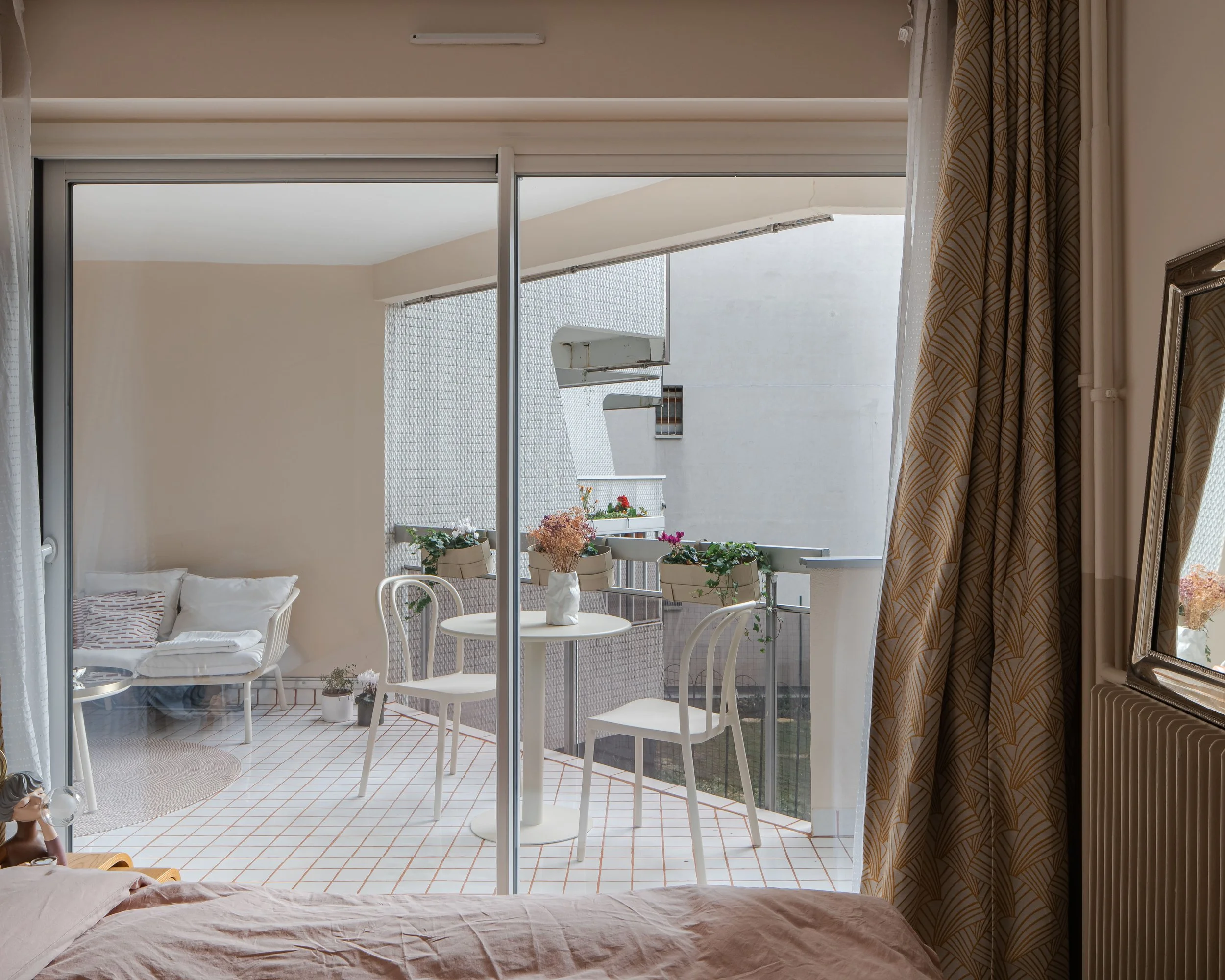 Balcon avec table, chaises et fleurs, vu depuis chambre avec fenêtre ouverte.