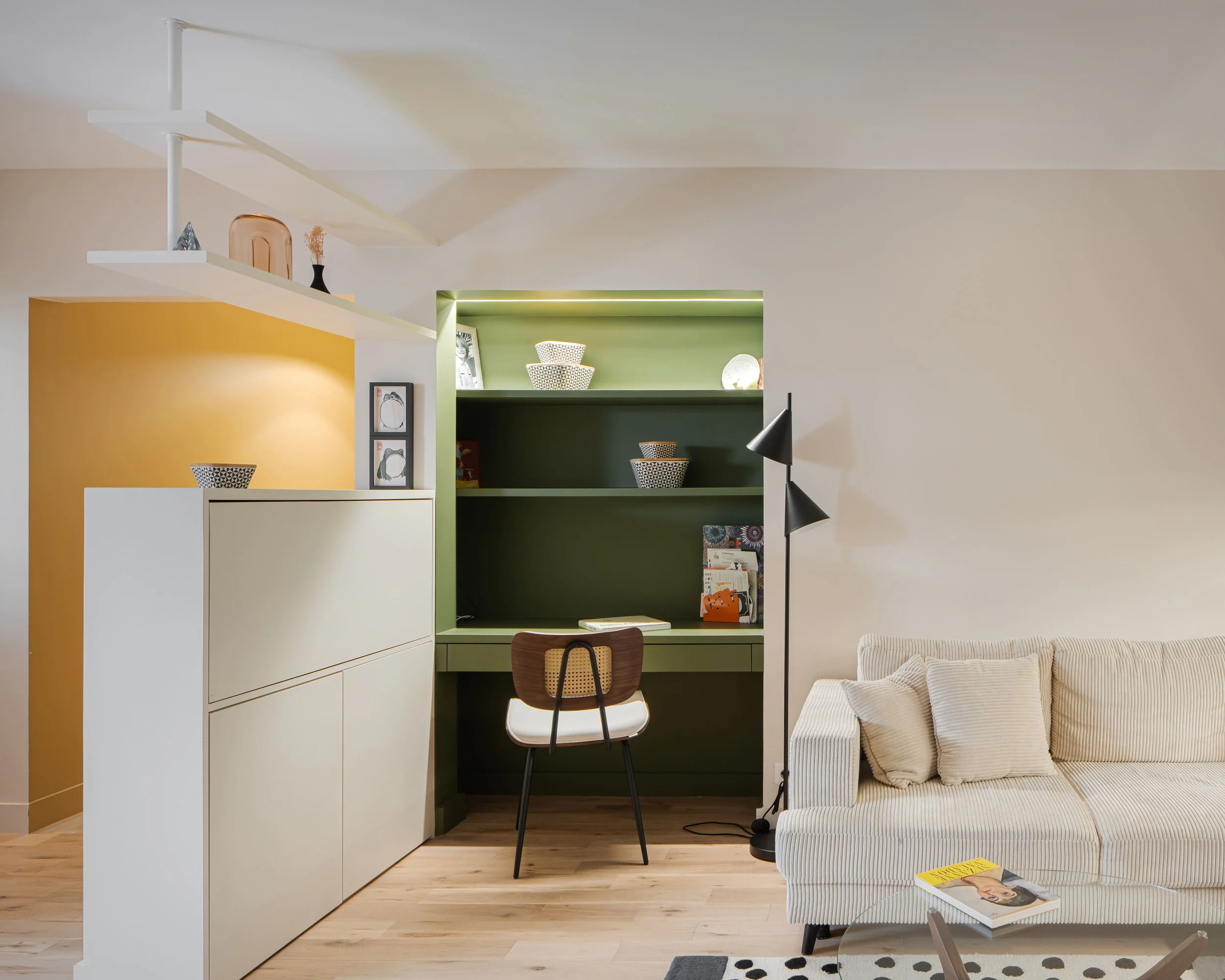 Intérieur moderne d’un salon avec étagères vertes, une lampe noire, un canapé beige rayé, une table avec un magazine, et un bureau en arrière-plan.