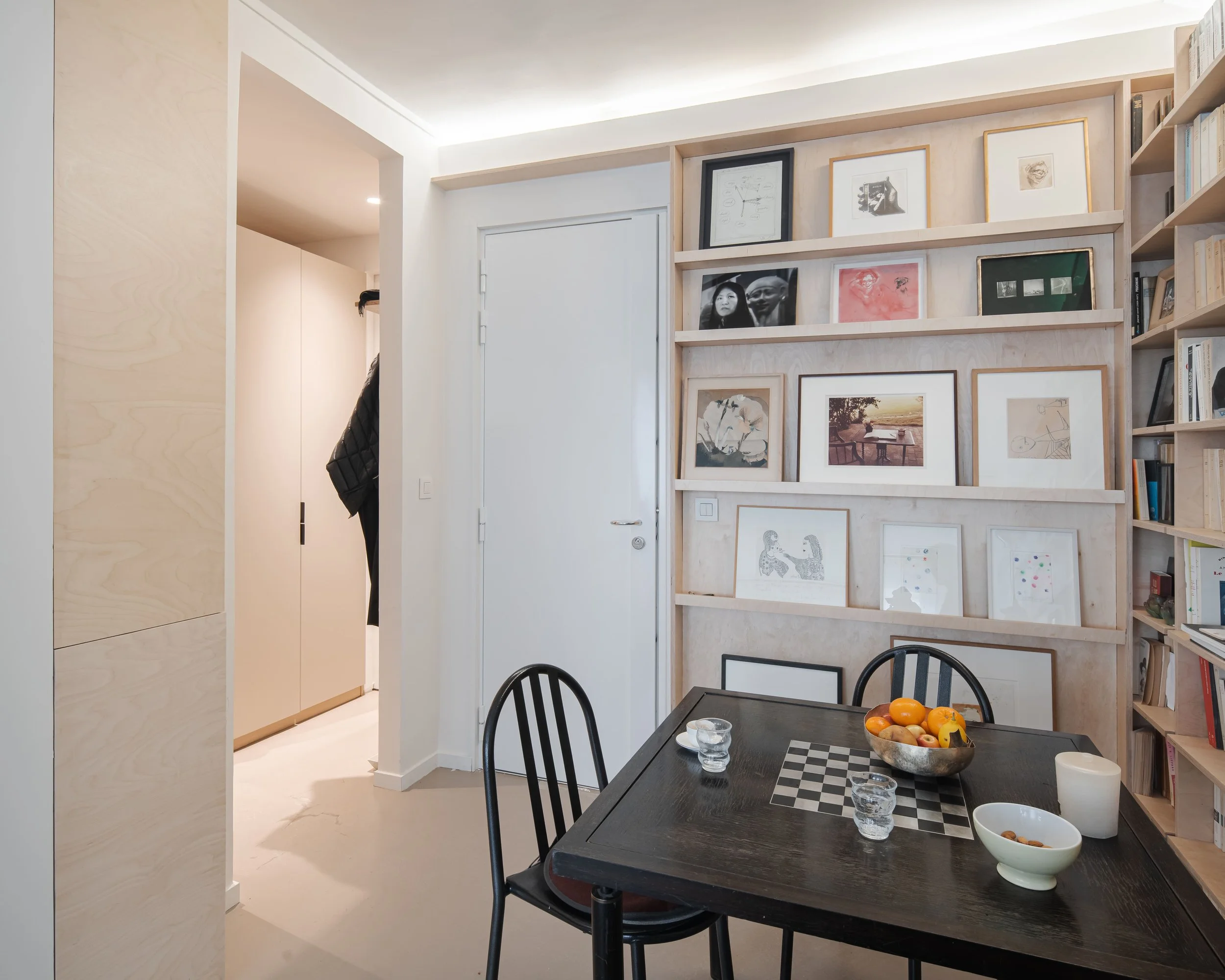 Intérieur moderne avec mur de cadres photos, table à manger avec fruits, livres et objets décoratifs, et porte d'entrée visible.