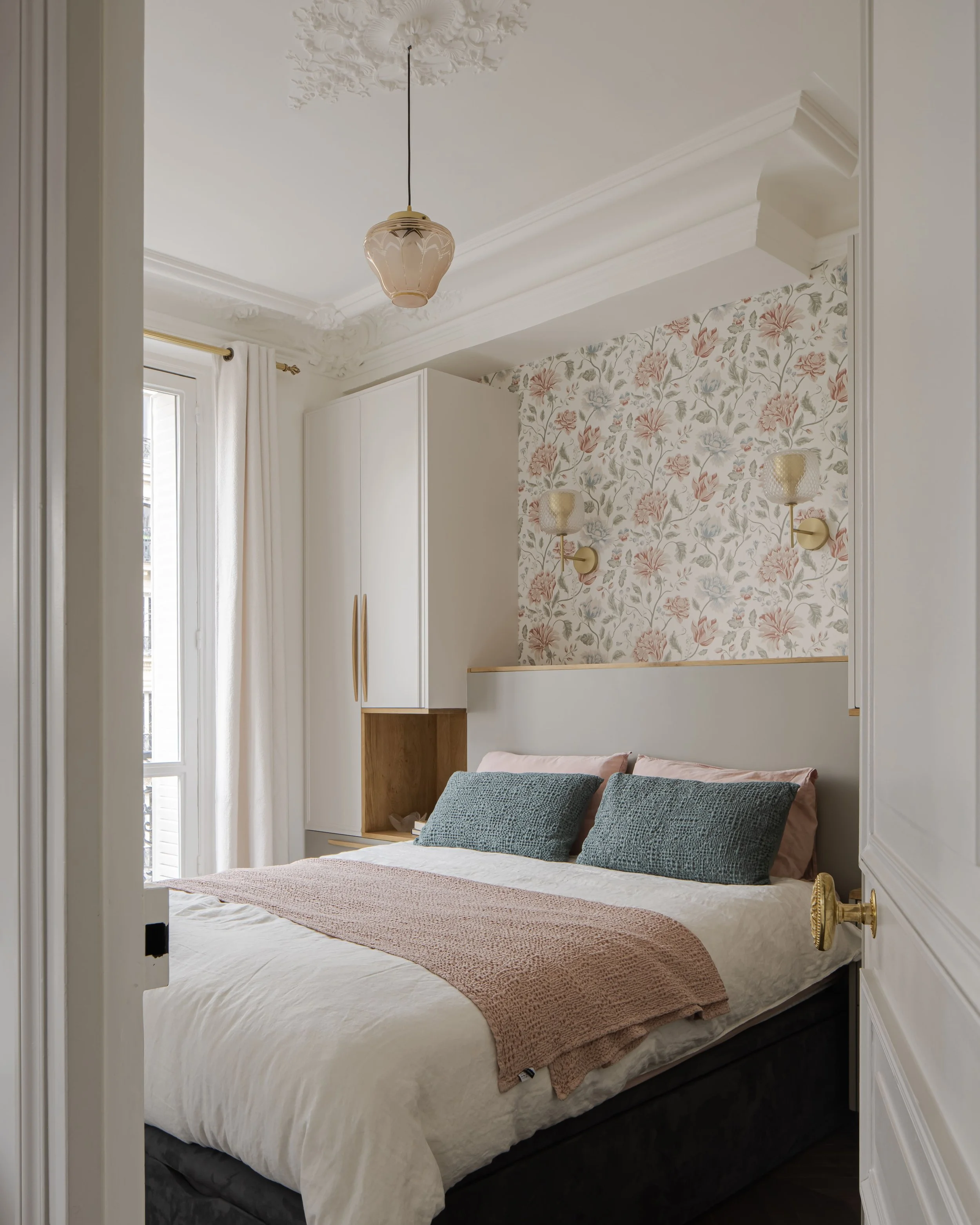 Chambre lumineuse avec lit, deux coussins gris, tête de lit, murs ornés de papier peint floral et appliques murales, fenêtre avec rideaux blancs et porte entrouverte.