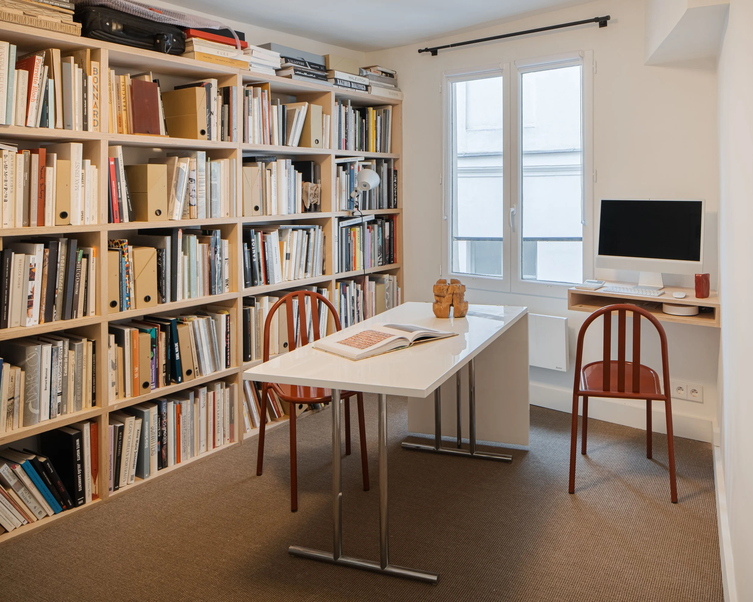 Une petite salle de lecture avec une grande bibliothèque remplie de livres, un bureau blanc avec deux chaises en bois, un ordinateur sur une étagère, deux fenêtres laissant entrer la lumière naturelle, et quelques objets décoratifs sur le bureau.