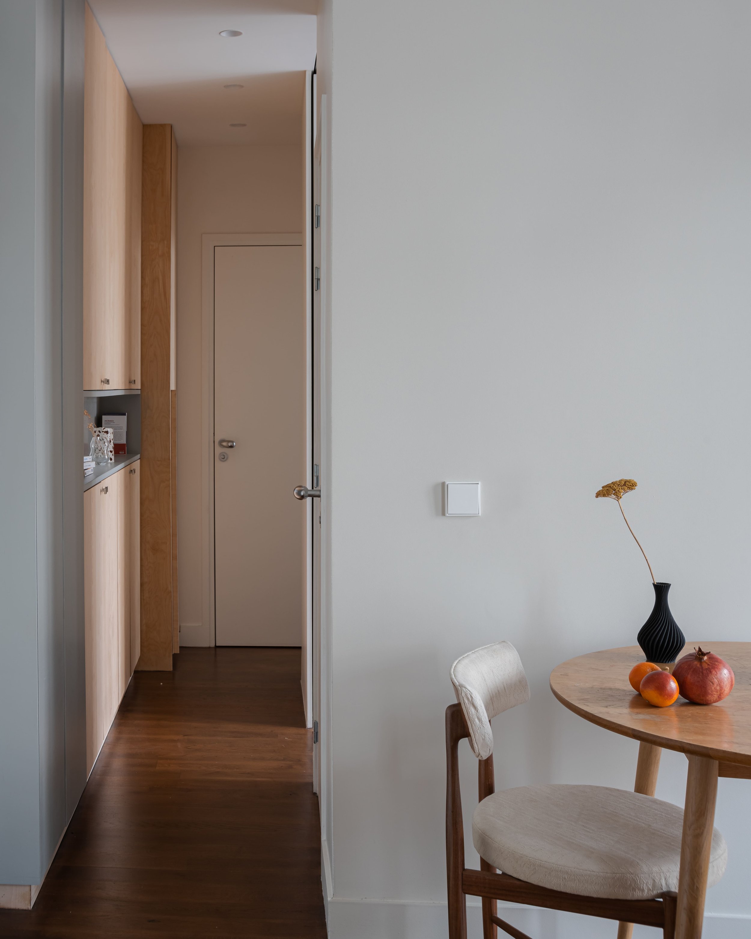 Une pièce intérieure avec un mur blanc, une table ronde en bois avec un vase noir et des fruits, et une chaise en bois et tissu beige. À gauche, un couloir avec un placard en bois clair et une porte blanche.