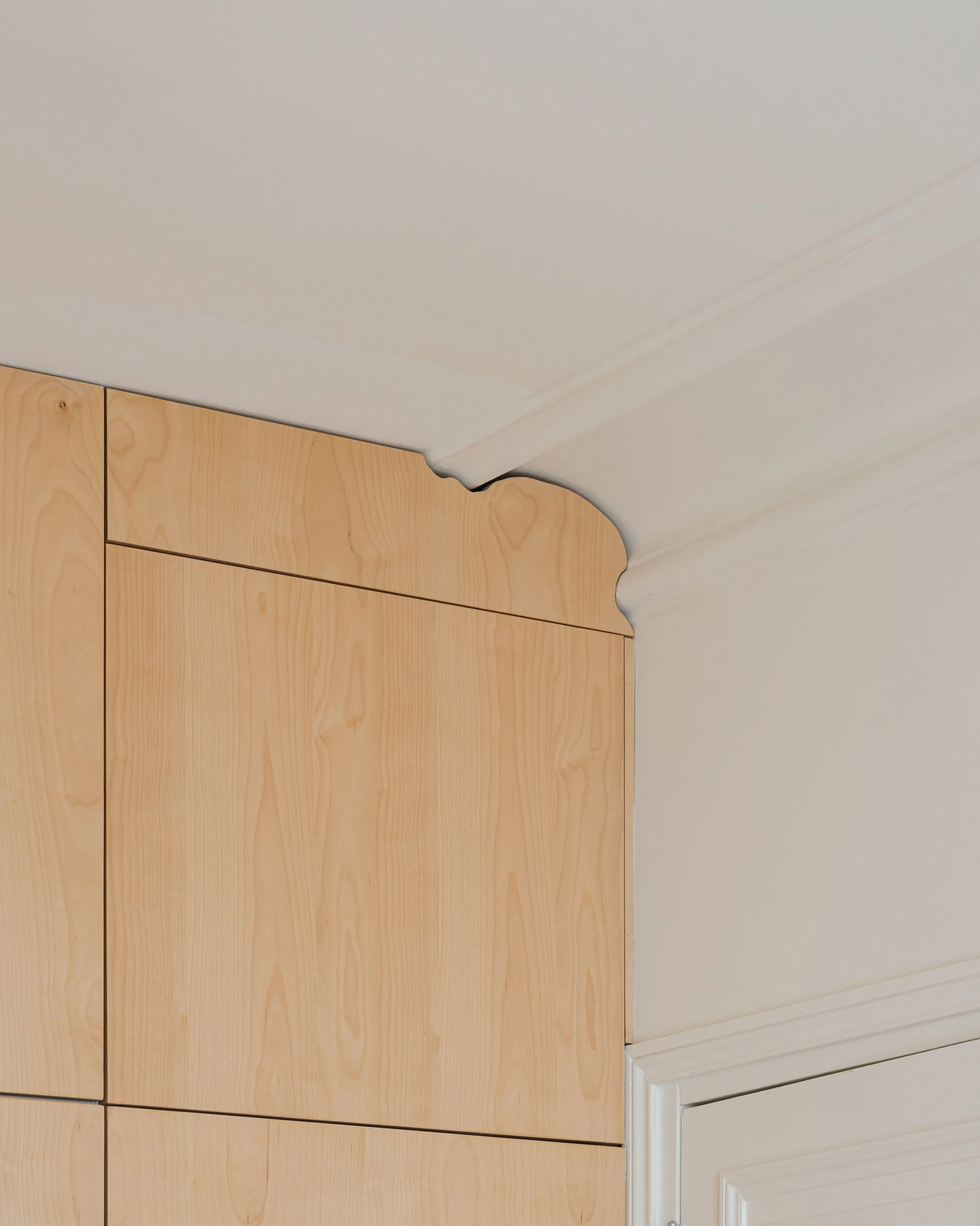 Détail d'une partie d'armoire en bois clair avec moulure en haut, située dans un intérieur avec mur blanc et porte blanche.