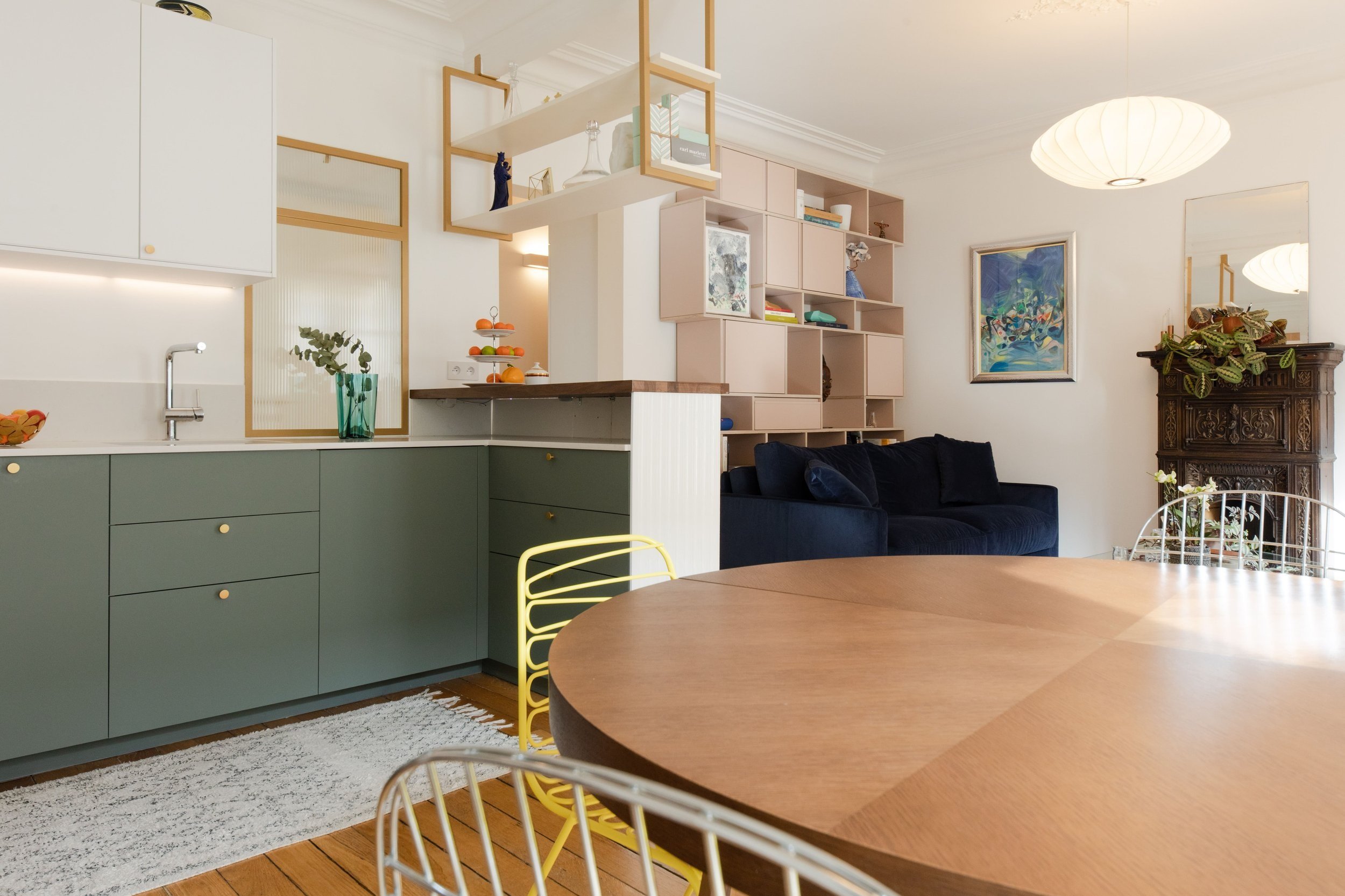 Intérieur d'un salon avec meuble de cuisine vert, canapé bleu, table ronde en bois, étagère décorative, peinture colorée et lampadaire blanc.