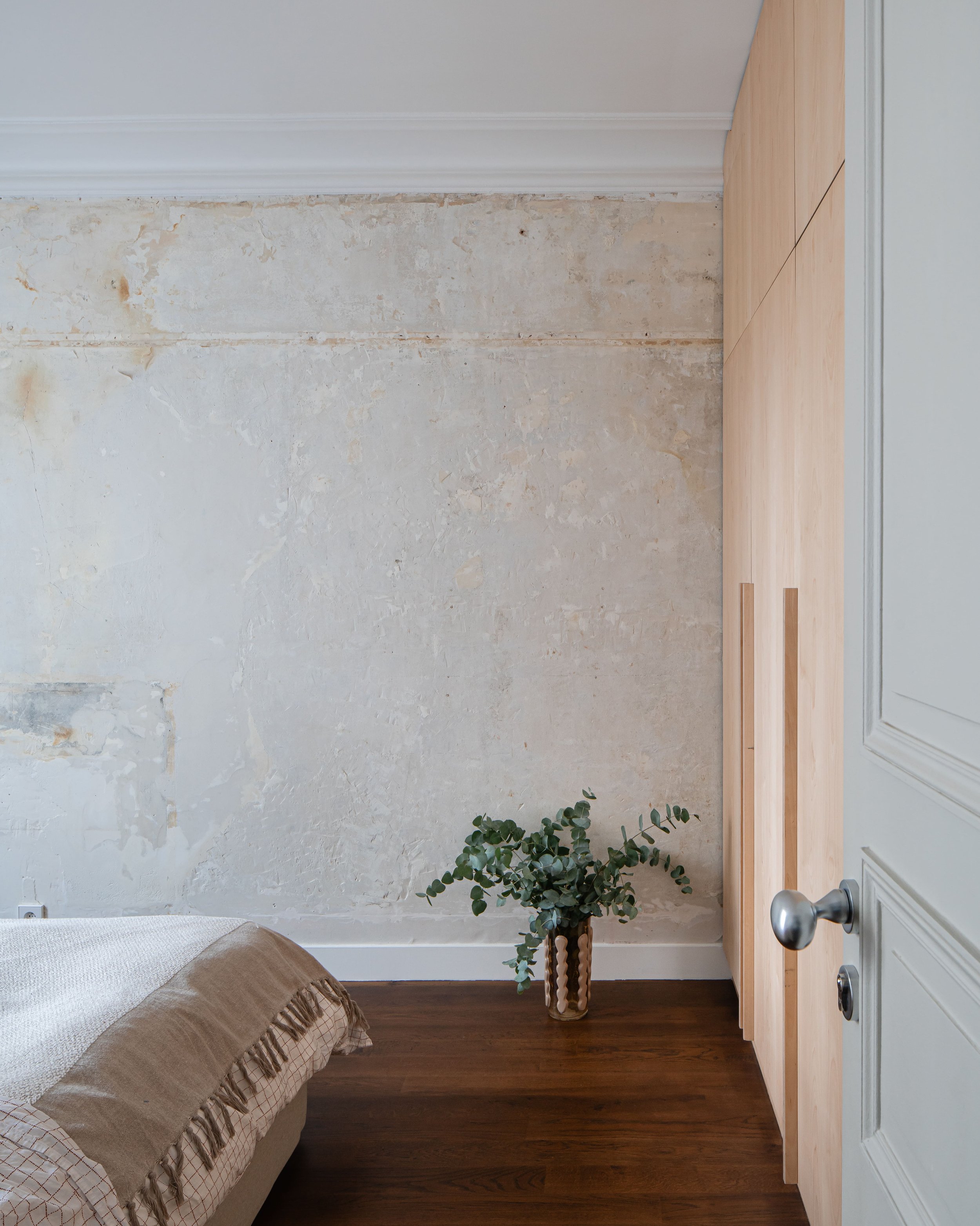 Chambre avec mur en pierre, lit avec couvre-lit beige, et une plante verte dans un vase, porte en bois clair, sol en bois foncé.