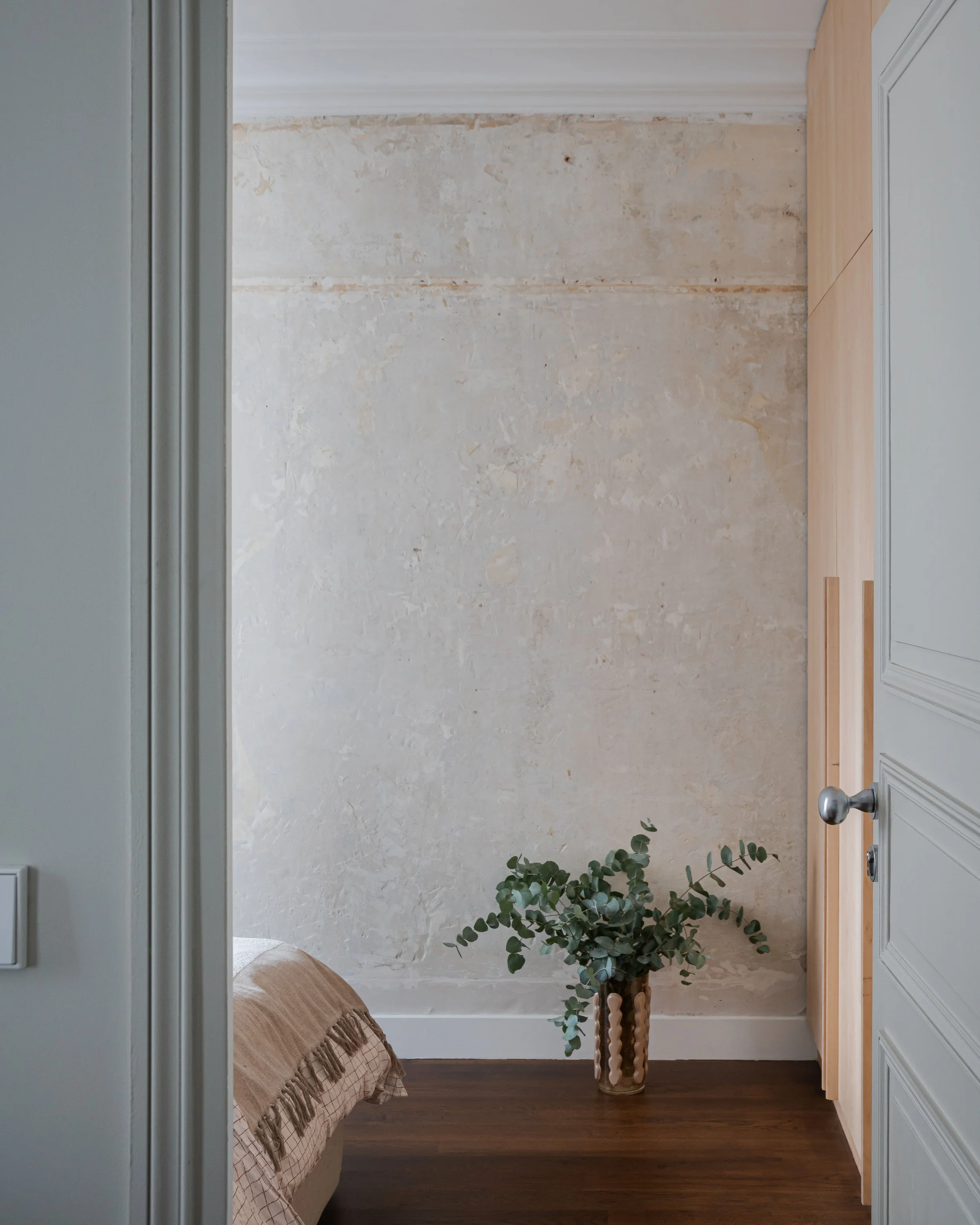 Une pièce avec un mur en plâtre, un sol en bois foncé, une plante verte dans un vase en verre, et un coin de lit avec une couverture beige.