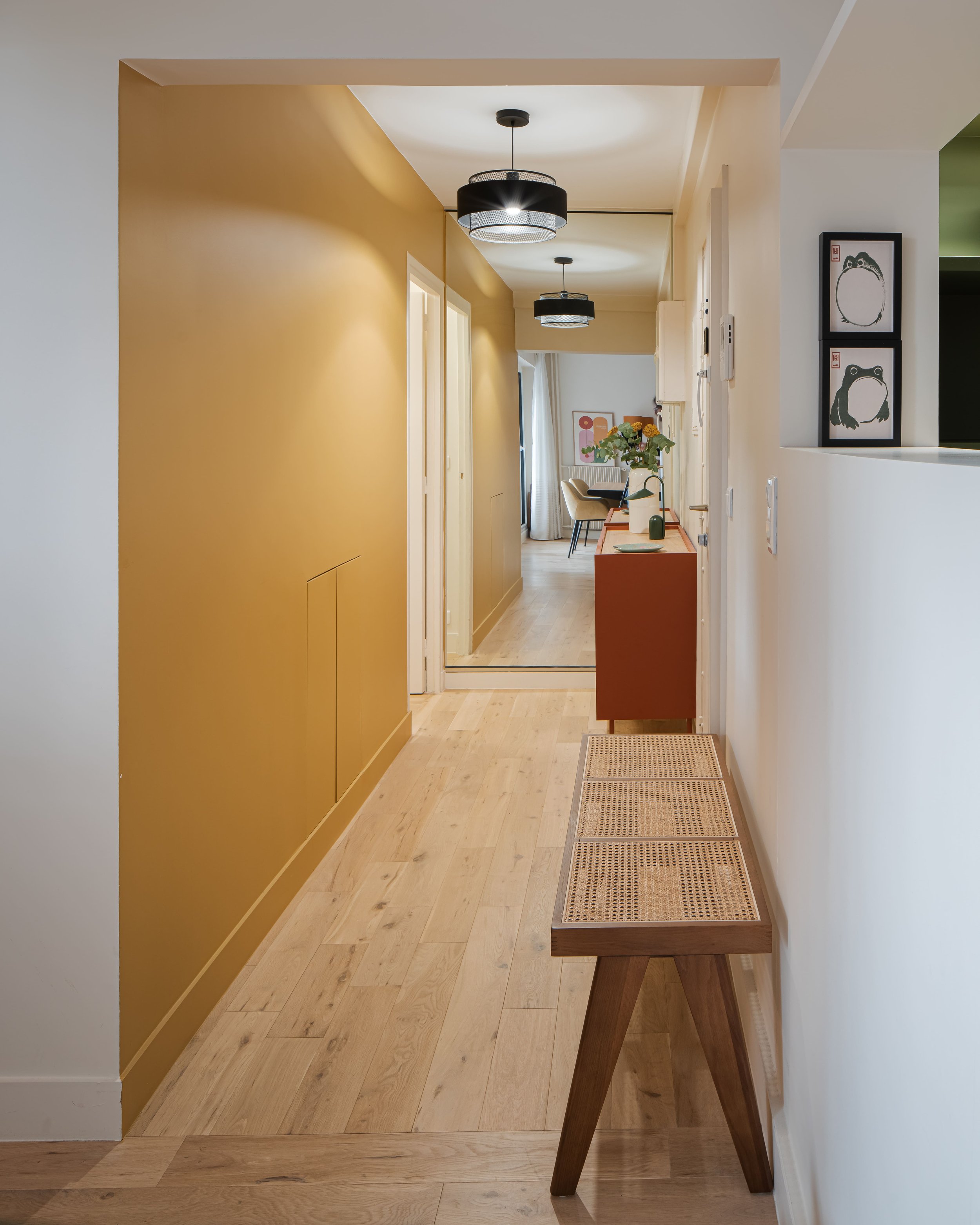 Un couloir intérieur avec un mur jaune sur la gauche, un parquet en bois clair, un meuble rouge avec des fleurs, et un mur blanc à droite avec deux cadres d'art mural. Des plafonniers noirs suspendus au plafond.