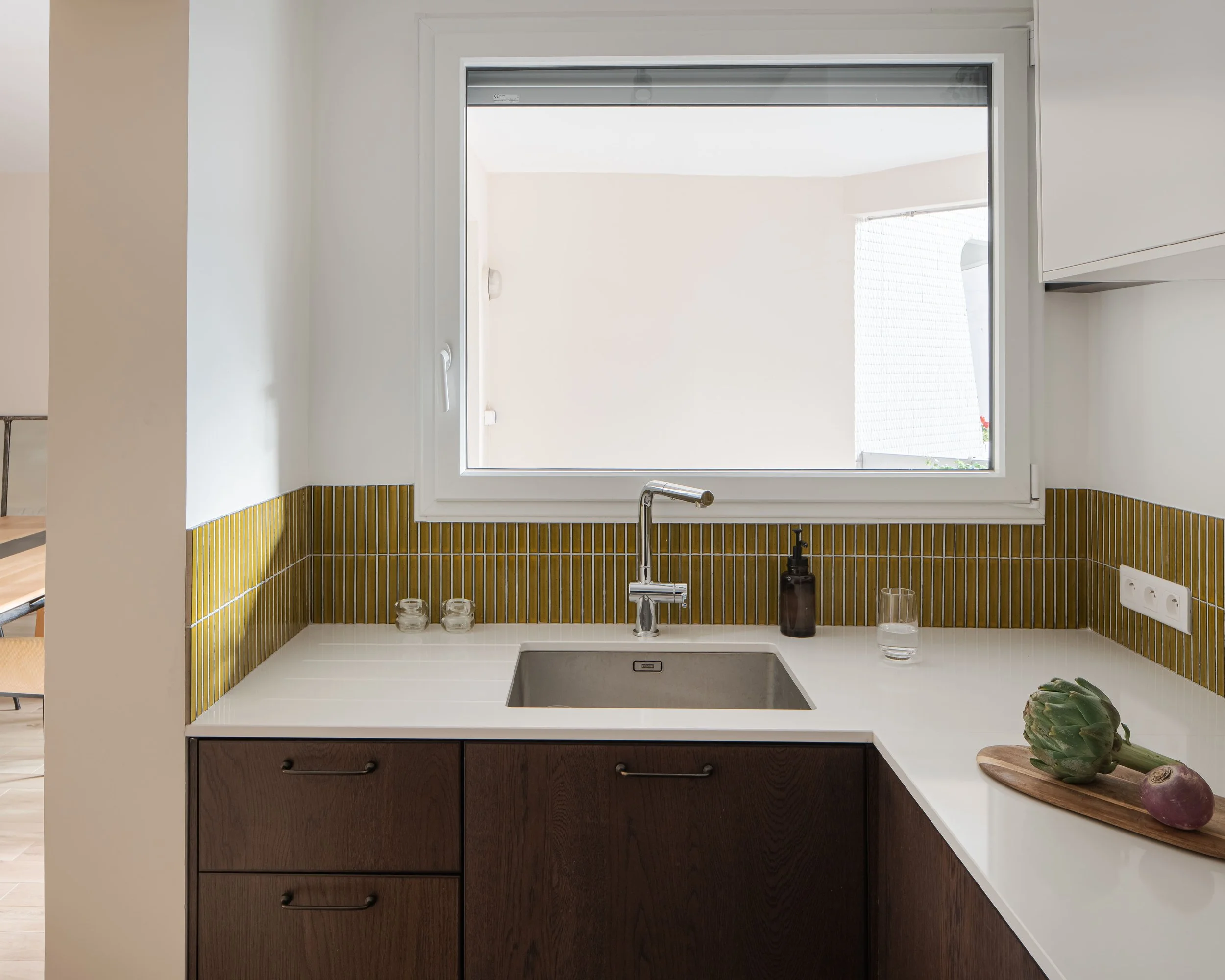 Une cuisine moderne avec un évier sous une fenêtre, une planche avec un artichaut et un oignon, des bocaux en verre, une bouteille en verre foncé, une prise électrique et un robinet.