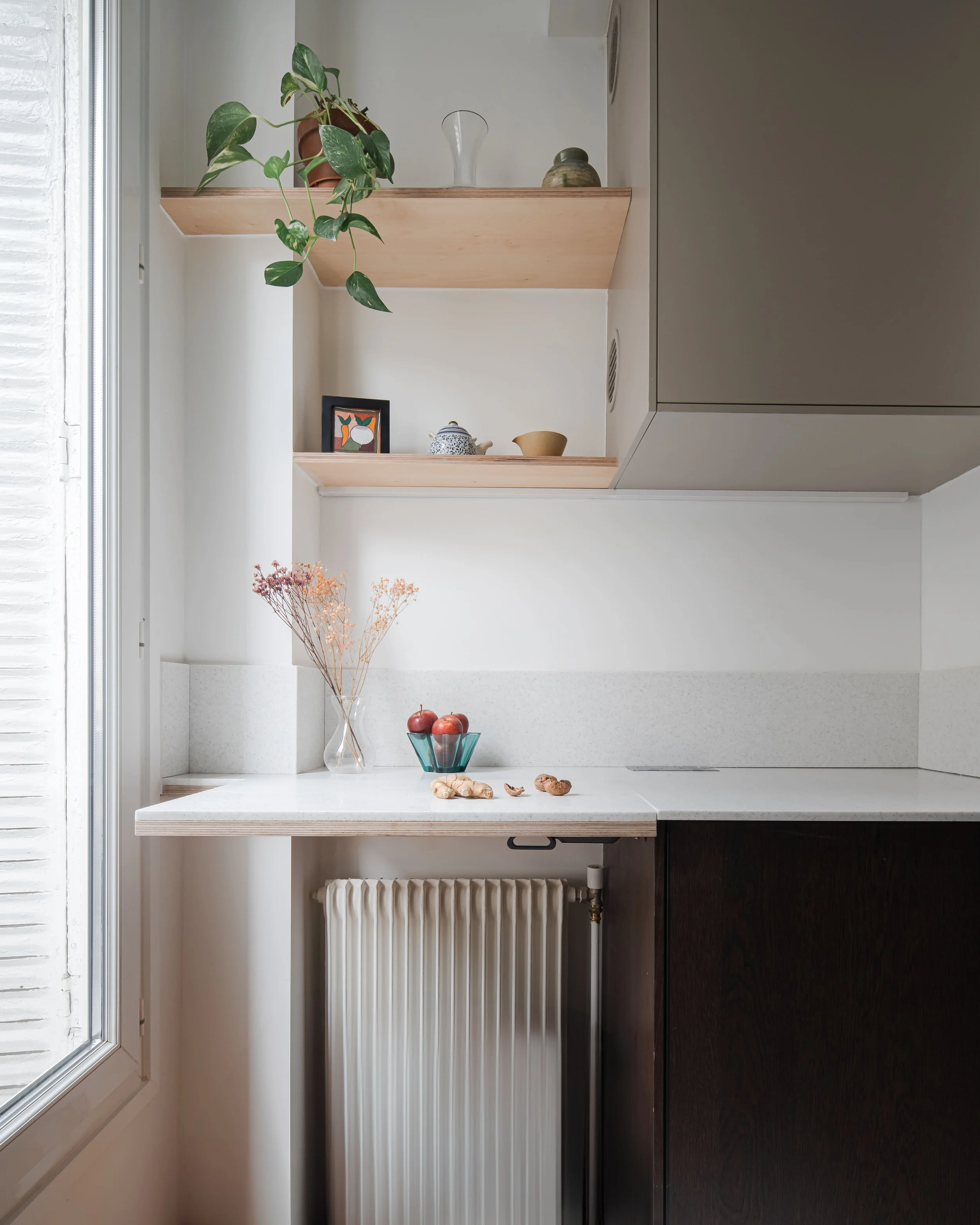 Coin de cuisine avec étagères en bois, vase avec fleurs séchées, bol de fruits, objets de décoration, et un radiateur blanc.