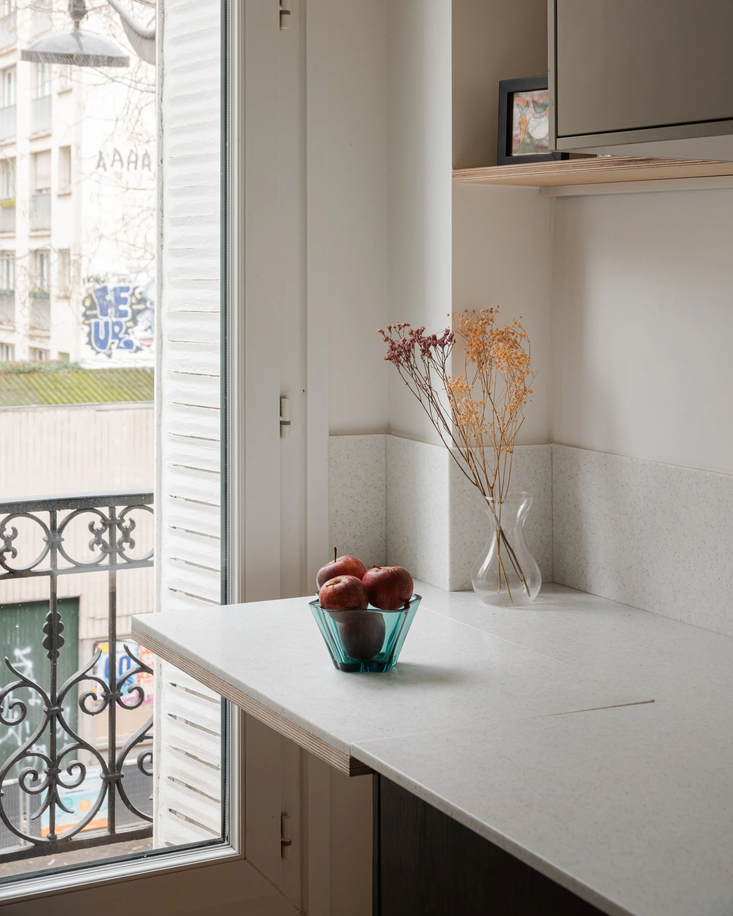 Une fenêtre ouverte donnant sur un balcon en fer forgé, avec un comptoir de cuisine blanc, un vase avec des fleurs séchées et un bol avec des pommes.
