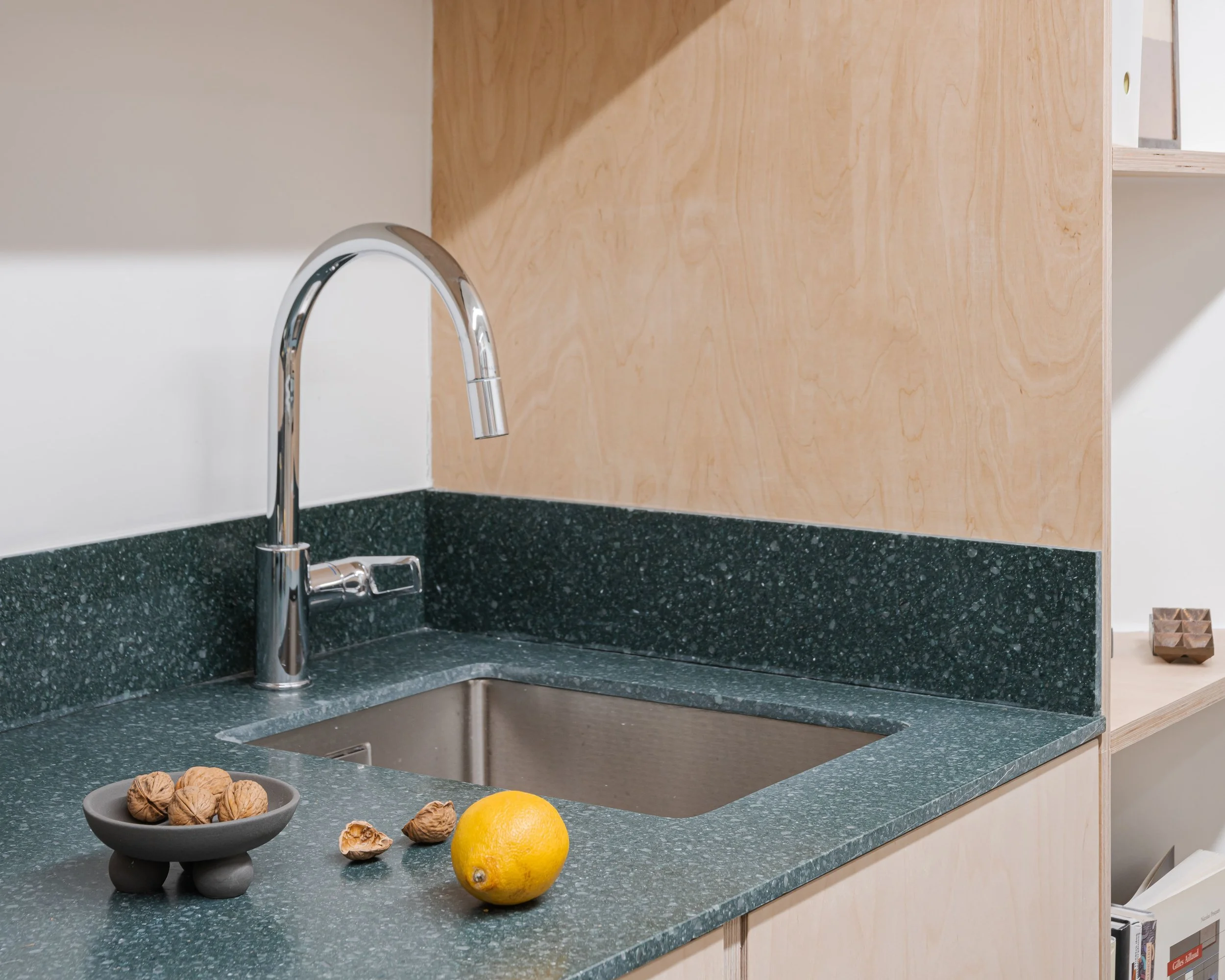 Coin de cuisine avec évier en acier inoxydable, robinet argenté, comptoir en pierre vert foncé, cier avec noix, citron et noix de coco, et bibliothèque en bois naturel montant vers le haut.
