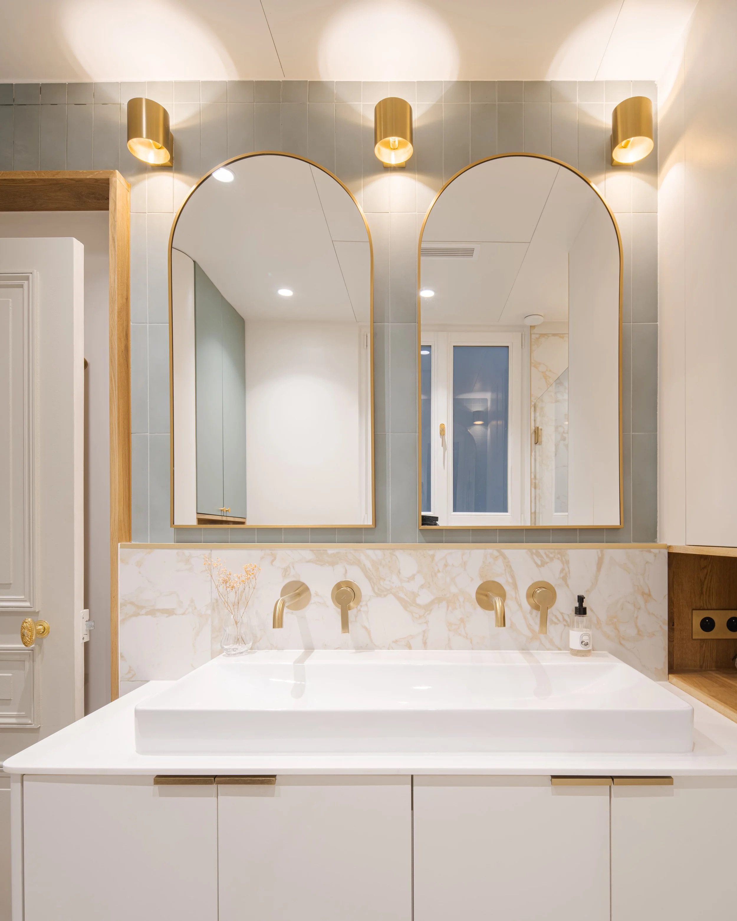 Miroir au-dessus d'un lavabo de salle de bain blanc avec espace de rangement en bois, appliques dorées, et fenêtre, intérieur moderne élégant.