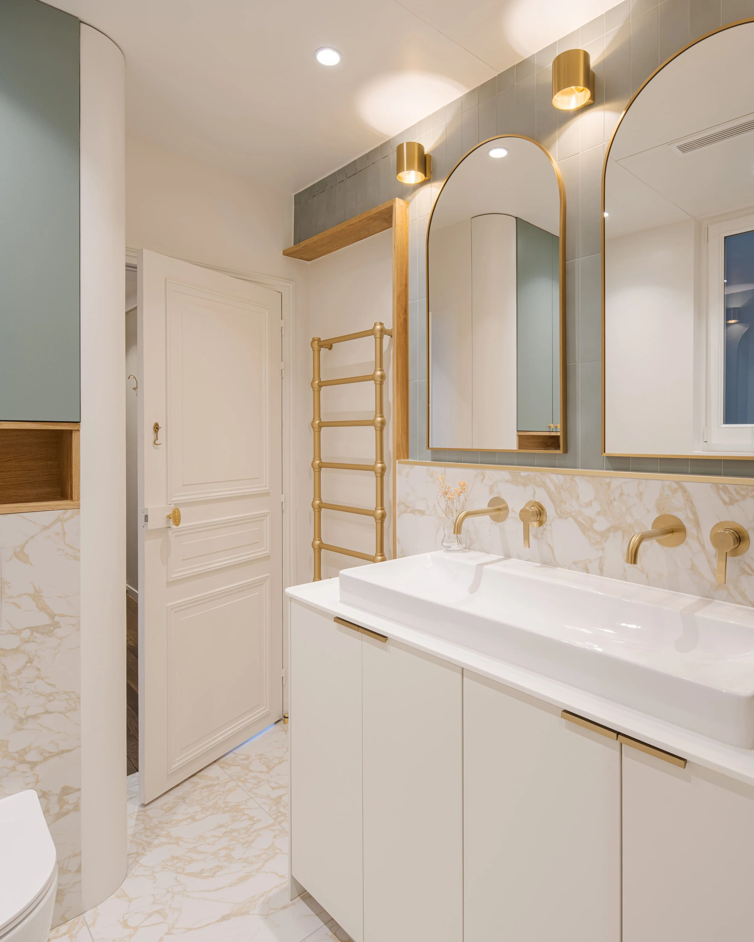 Salle de bain moderne avec un miroir, un lavabo blanc, et des robinets dorés, murs avec du carrelage brillant, éclairage chaud, porte blanche, et éléments en bois et doré.