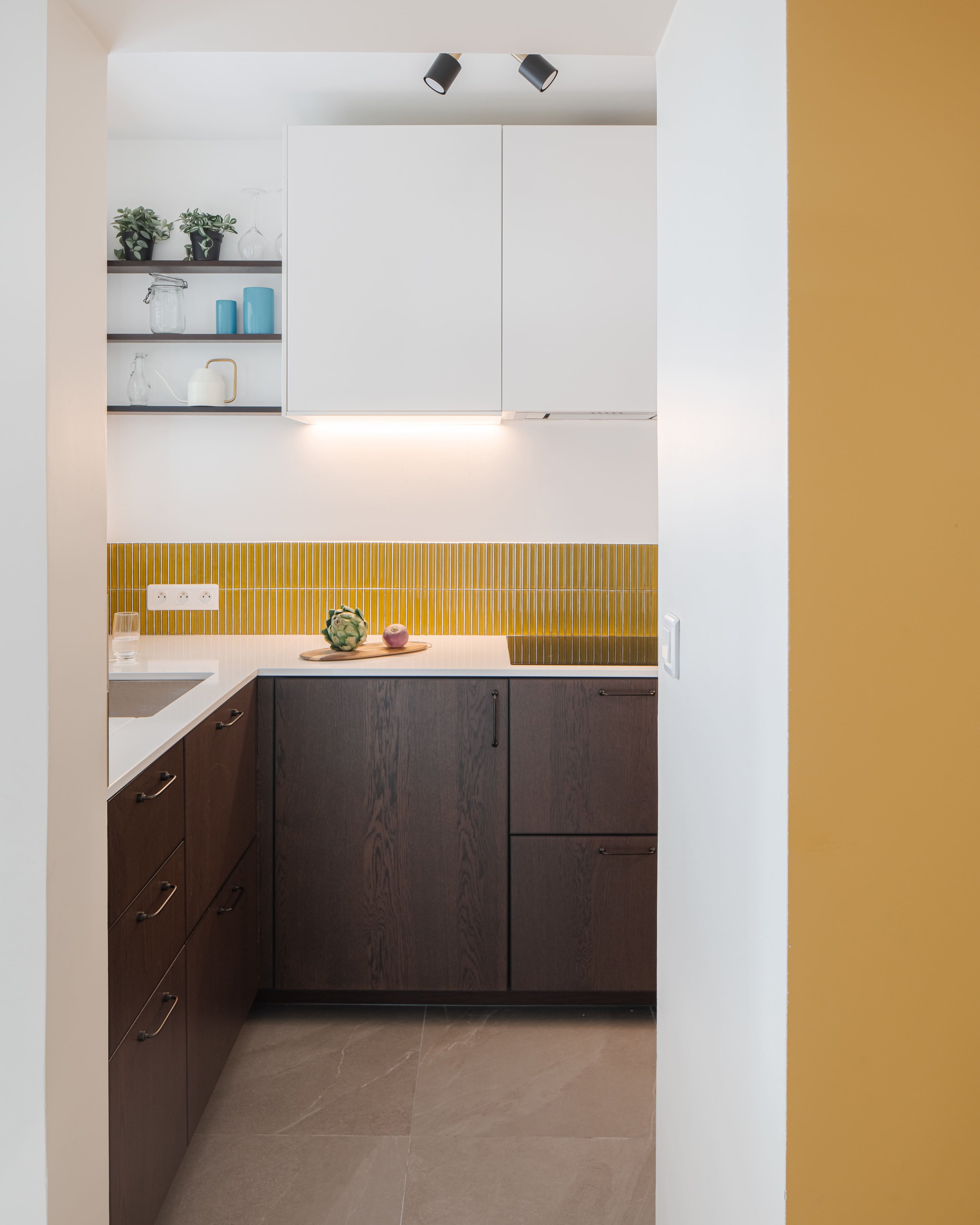 Petite cuisine moderne avec des armoires en bois foncé, un plan de travail blanc, un mur de carrelage jaune, des étagères avec des objets en verre, plates et des plantes, une lumière douce sous les armoires et un plafonnier à deux spots