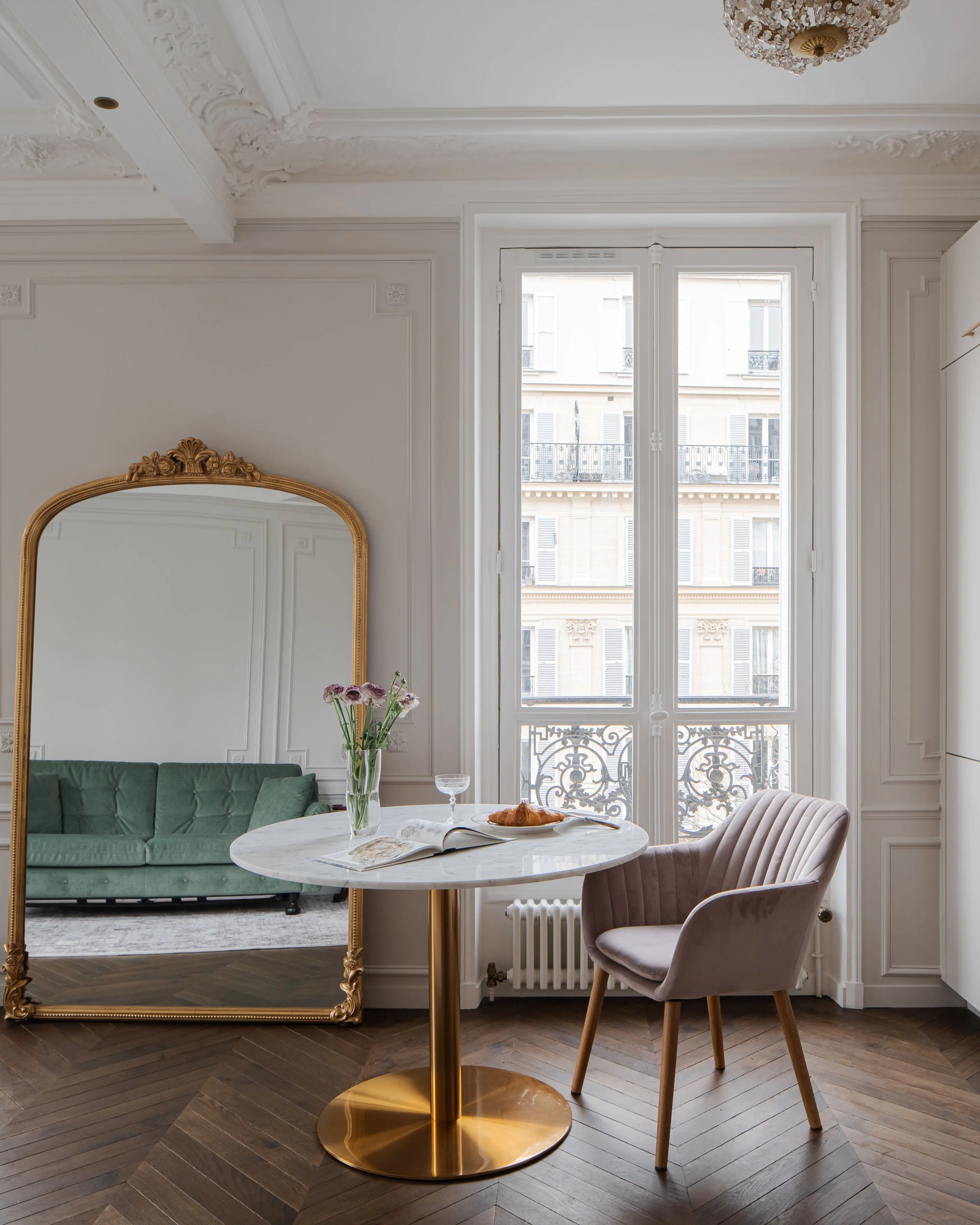 Intérieur d'un appartement élégamment décoré avec une grande fenêtre, un miroir ancien doré, une table ronde en marbre avec un vase de fleurs, un canapé vert, une chaise en velours rose pâle, une lampe en plâtre, et un parquet en bois.