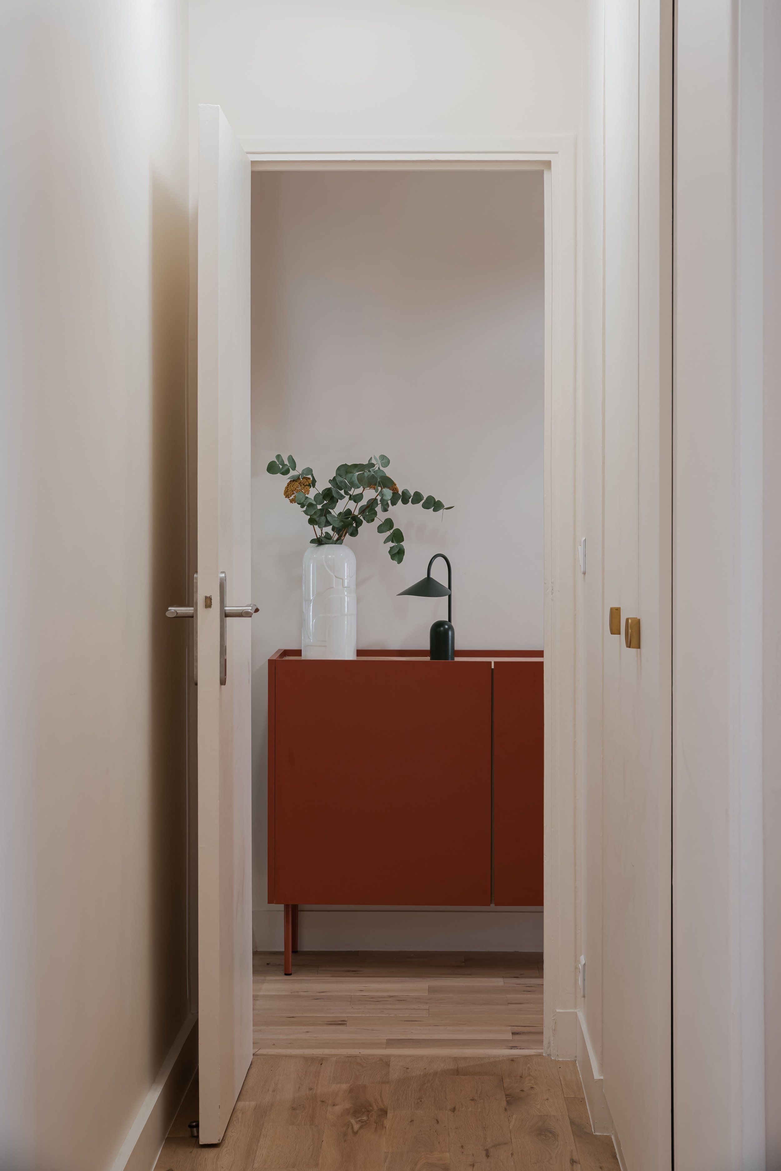 Vue d’un couloir menant à un meuble rouge avec un vase blanche contenant des branches de feuilles vertes et une lampe noire