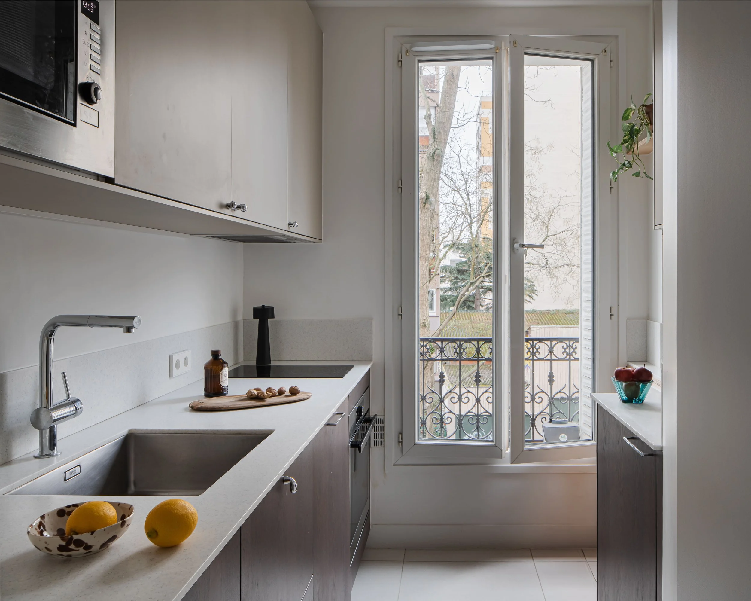 Cuisine moderne avec fenêtre donnant sur un balcon et des arbres, comptoir avec fruits et épices, armoires blanc et bois clair, accessoires de cuisine.