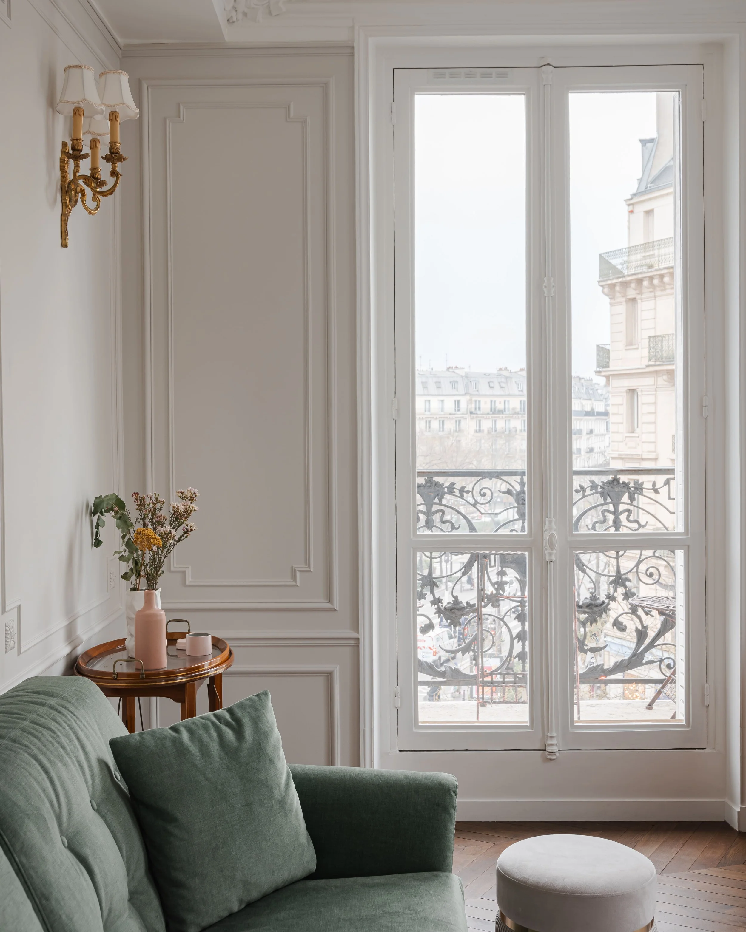 Intérieur d'un salon avec un canapé vert, une petite table en bois avec un vase à fleurs et deux bougies, une grande fenêtre avec un balcon en fer forgé et une vue sur des bâtiments parisiens.