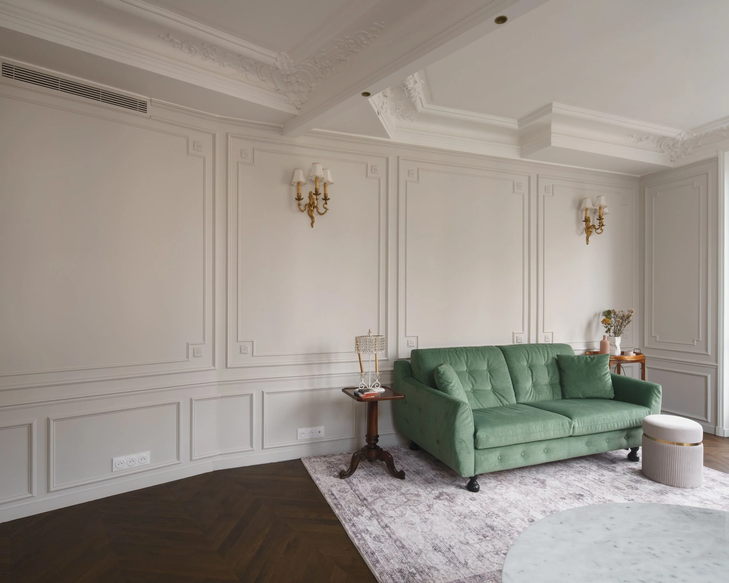Salon élégant avec murs blancs, canapé vert, tapis, tables d'appoint en bois, moulures décoratives au plafond, appliques murales dorées.
