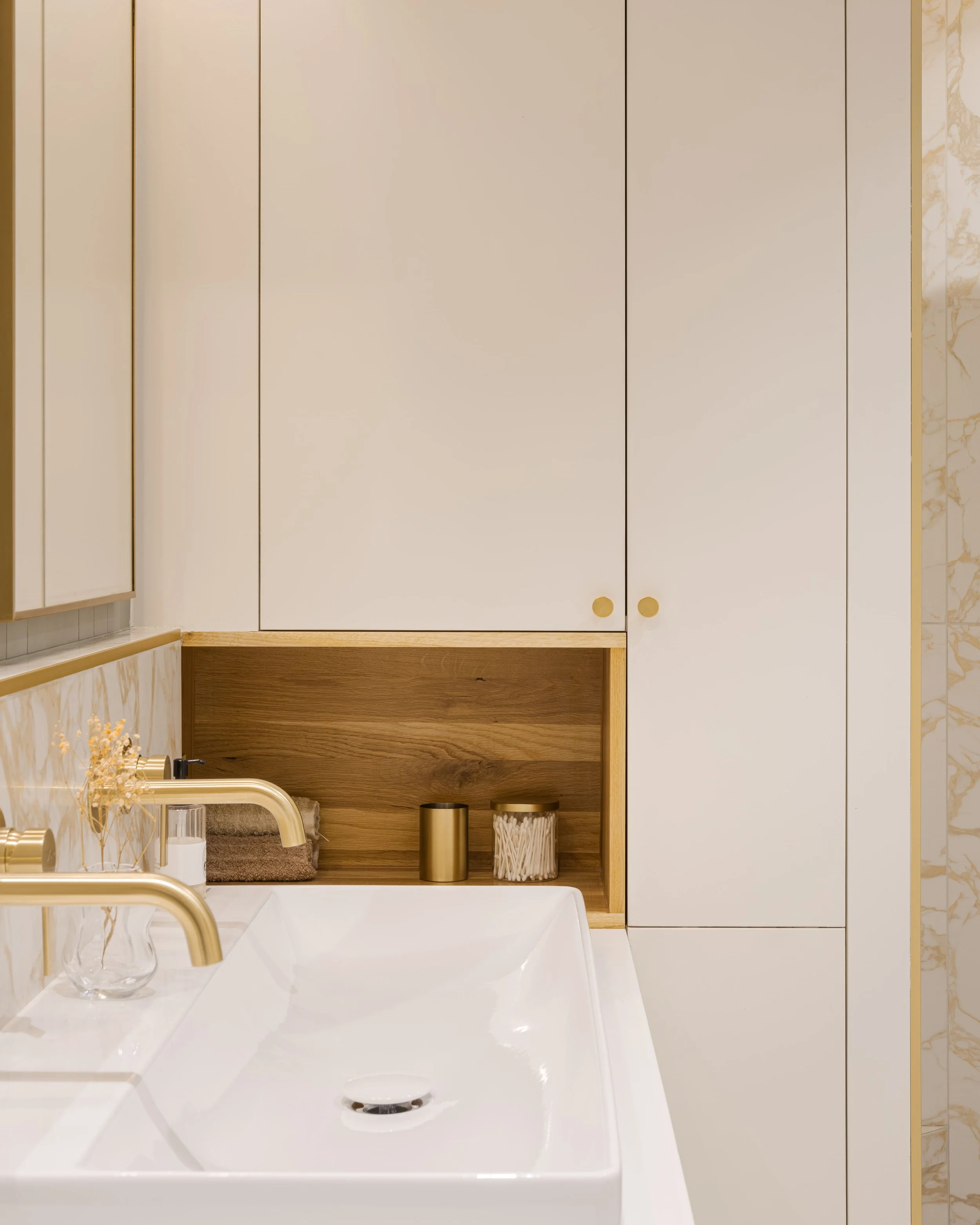 Vue proche d'un lavabo dans une salle de bain élégante avec des meubles blancs et des accents en bois, comprenant un vase avec des fleurs, deux récipients, et un robinet doré.