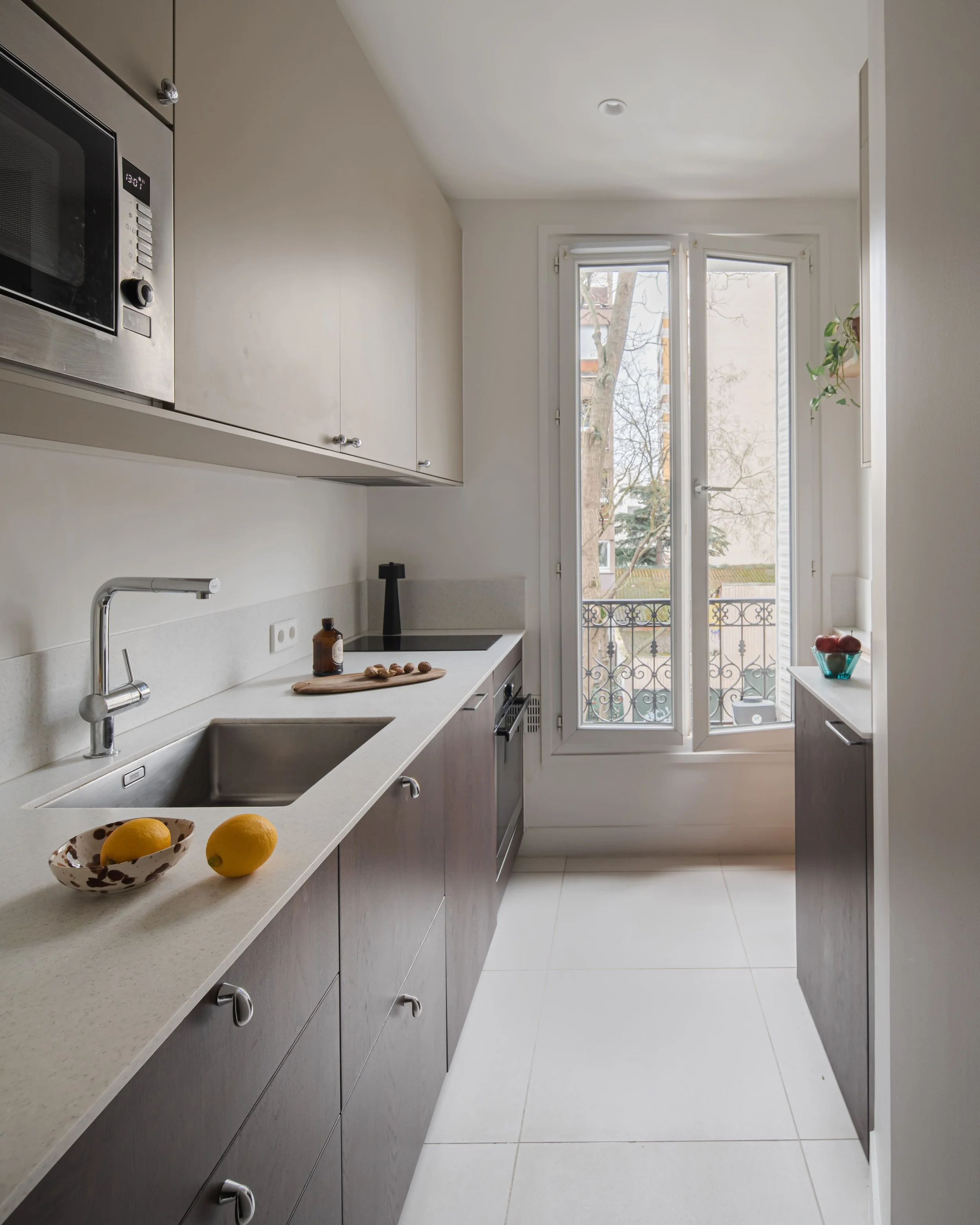 Cuisine moderne avec comptoir en quartz, évier, micro-ondes, fenêtres laissant entrer la lumière naturelle, décor minimaliste avec quelques fruits et objets.